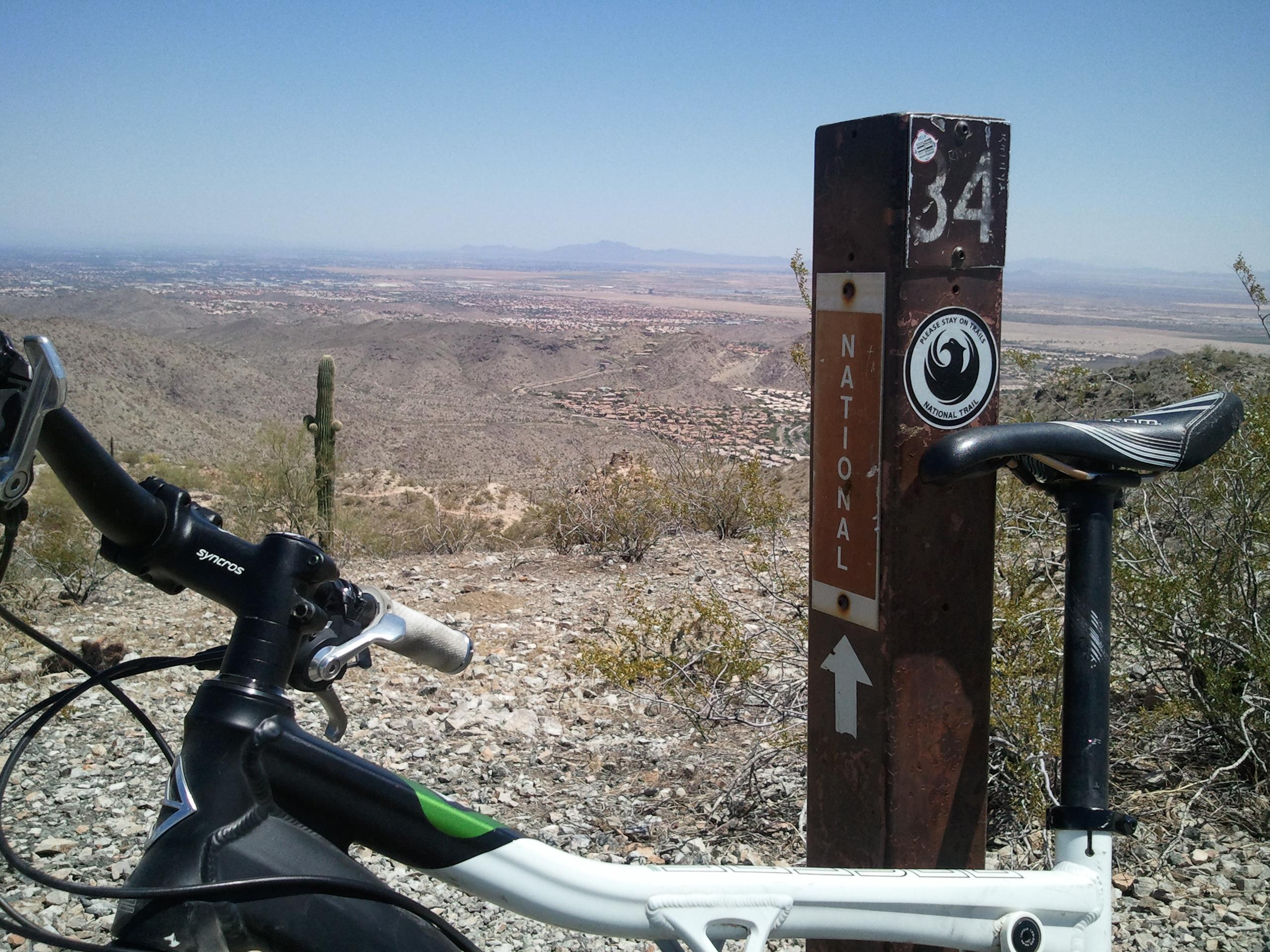 Gary Fisher Roscoe: A mountain bike is positioned in the foreground, leaning against a trail marker post labeled "34" with the word "NATIONAL" above and an arrow pointing upward. The background features a panoramic view of arid mountains and valleys under a clear blue sky. Cacti and sparse vegetation are visible in the surrounding area, indicating a desert landscape.