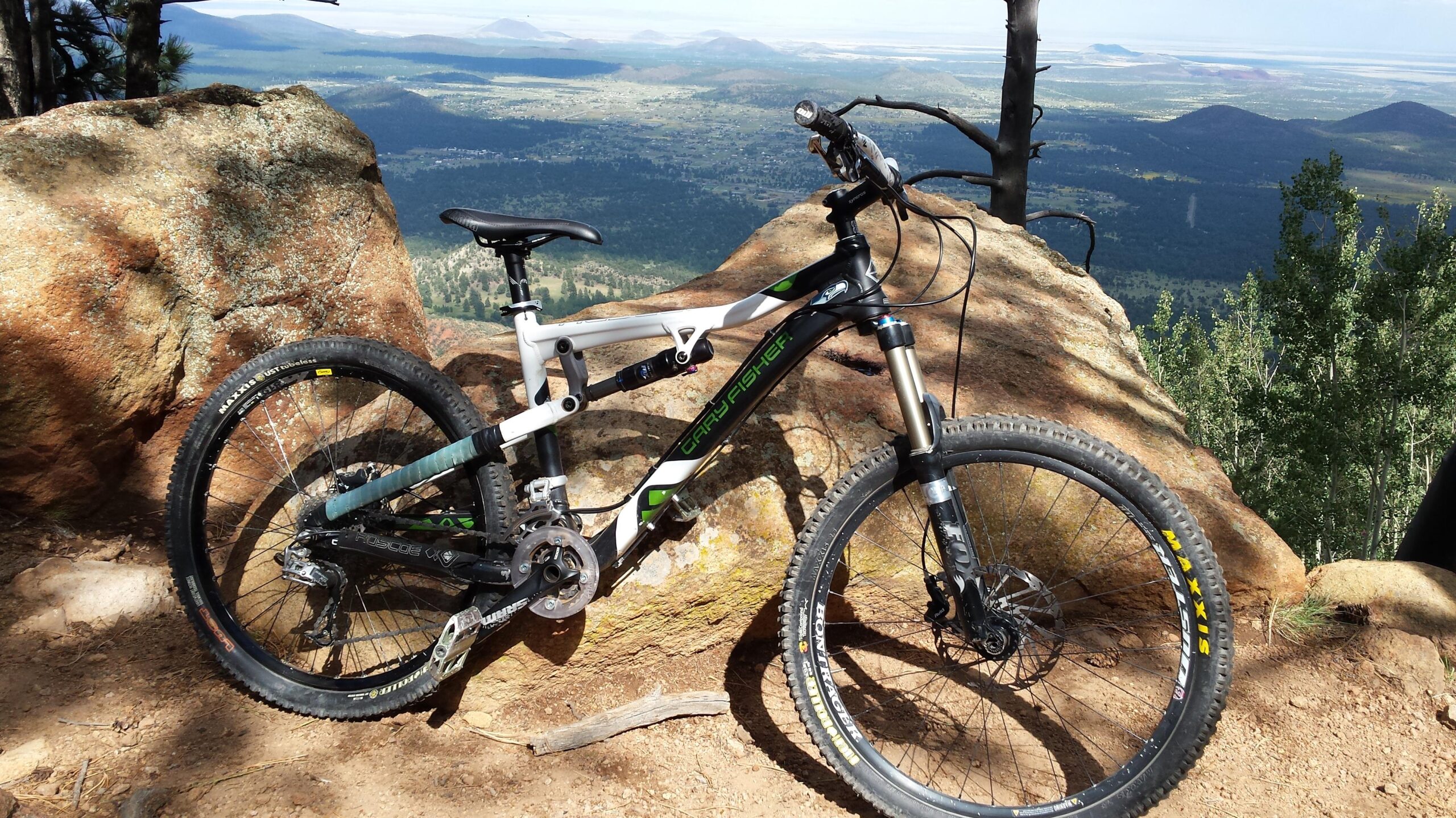 Gary Fisher Roscoe: A mountain bike rests against a large rock on a scenic overlook, with a panoramic view of green hills and distant mountains in the background. The sun casts shadows, highlighting the bike's frame and tires on the dirt path.