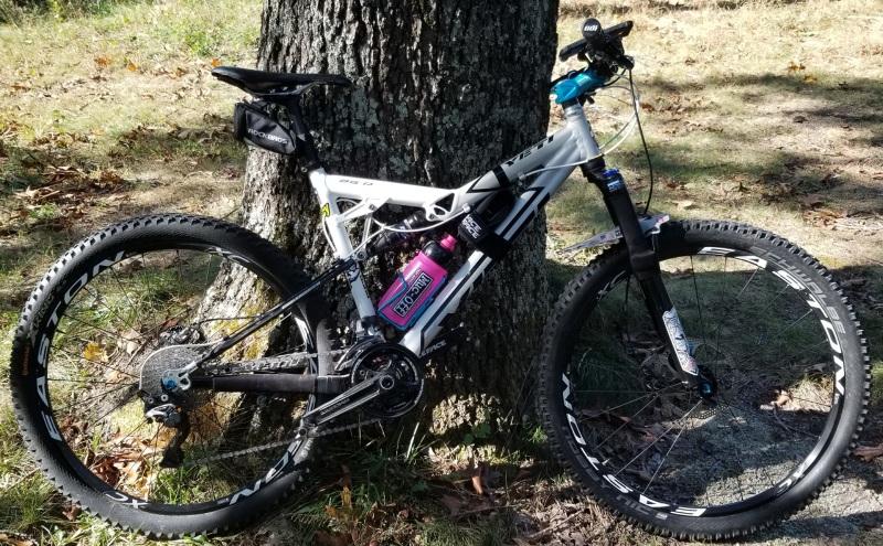 Yeti ASR - SLC: A mountain bike with a white frame and black tires is leaning against a tree in a natural setting. The bike features a pink water bottle and various components indicating it is designed for rugged terrain. The ground is covered in grass and leaves, suggesting an outdoor trail.