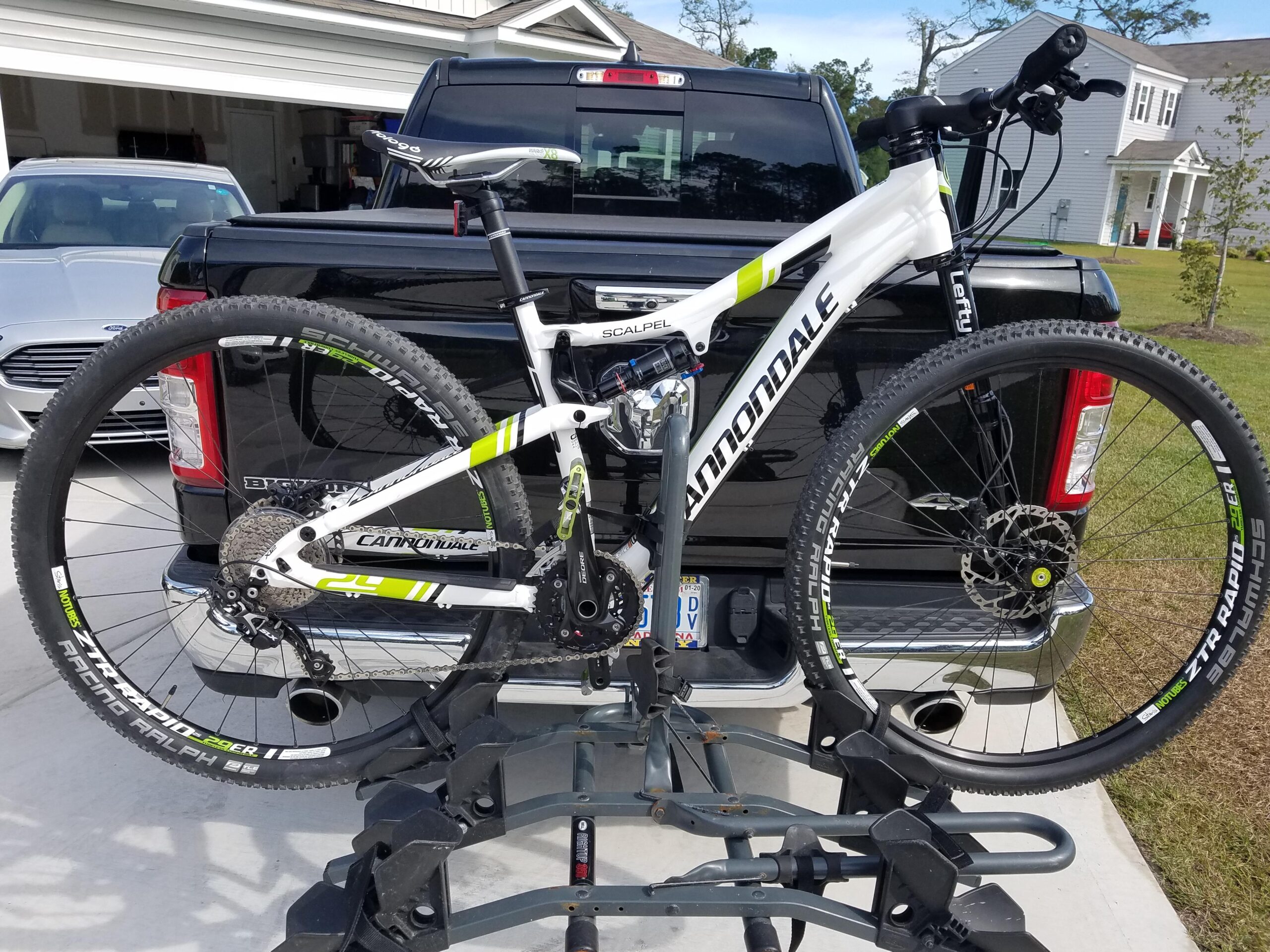 Cannondale Scalpel 29er 3: A white Cannondale Scalpel mountain bike mounted on a bike rack attached to a black pickup truck. The bike features large tires, a modern frame design, and is equipped with a Lefty front suspension fork. In the background, a silver car can be seen parked in a driveway next to a house.