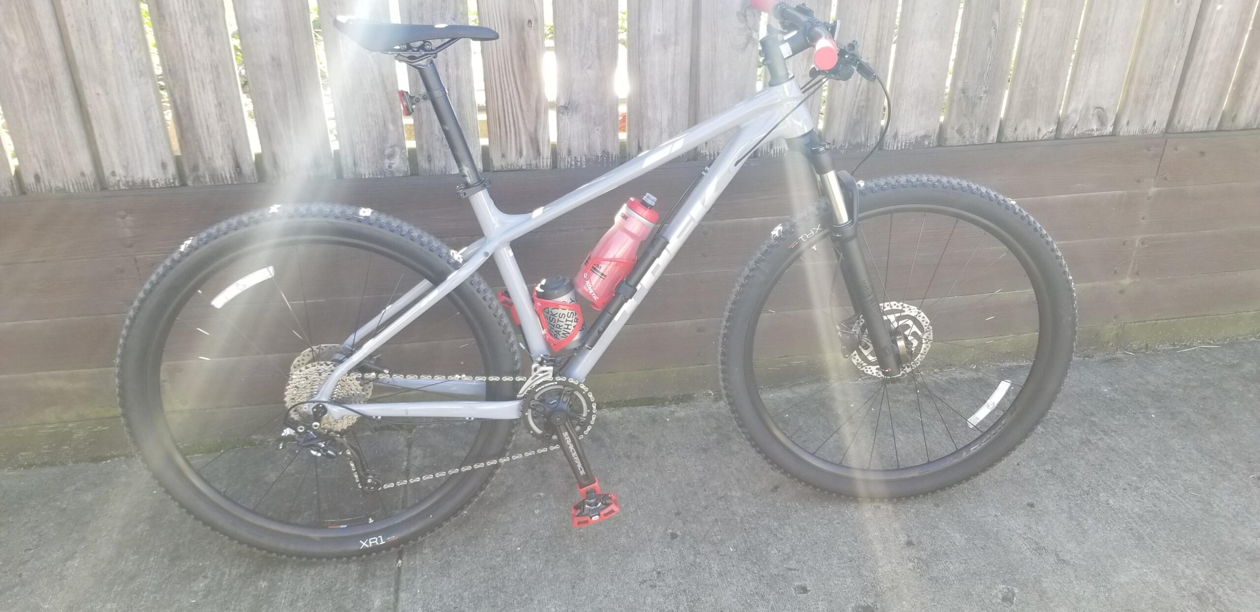Trek X-Caliber 8: A gray mountain bike leaning against a wooden fence, featuring a prominent suspension fork, thick tires, and a water bottle holder with a red water bottle. The bike has a 1x drivetrain setup and visible chain components. Sunlight is illuminating the scene, creating a bright and inviting atmosphere.