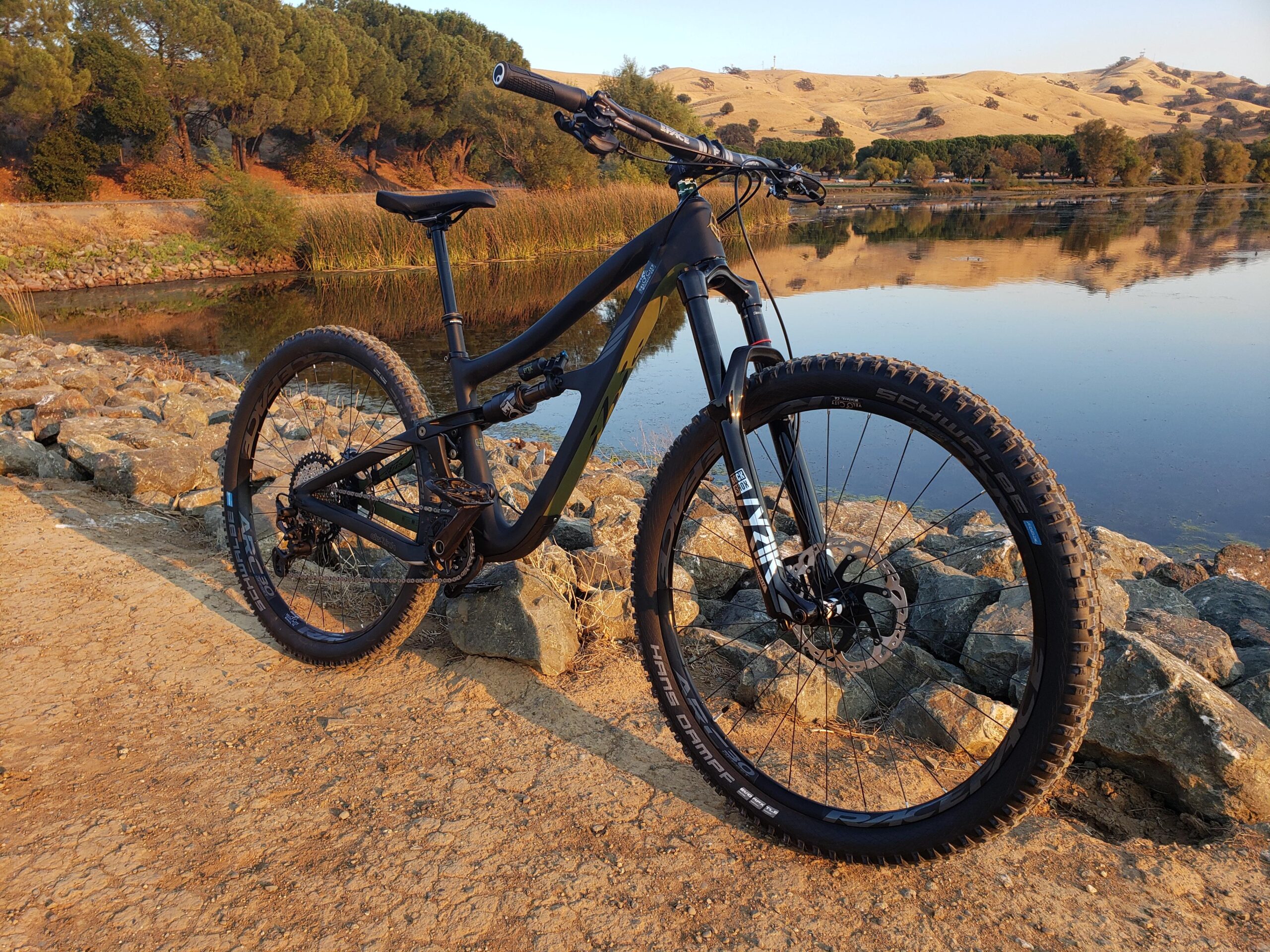 Ibis RipMo: A mountain bike resting on rocky terrain next to a body of water, surrounded by lush greenery and hills in the background. The setting is illuminated by warm, golden light, suggesting either early morning or late afternoon.