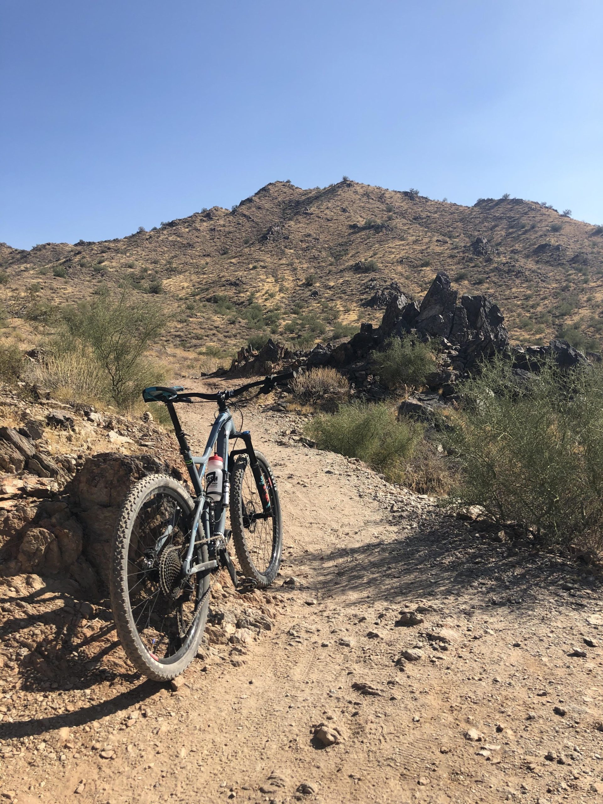 A mountain bike leaning against a rocky outcrop on a dirt trail, with rolling hills and a clear blue sky in the background. The landscape features sparse vegetation, typical of a dry, arid environment. San Tan mountain bike trail.