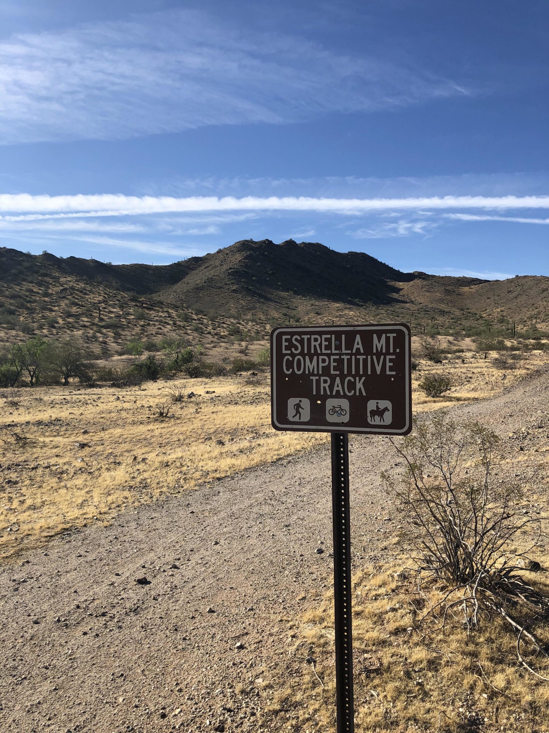 Sign for Estrella Mountain Competitive Track, indicating trails for hiking, biking, and horseback riding, with a backdrop of arid hills and a blue sky streaked with clouds. Estrella Mountain Park mountain bike trail.