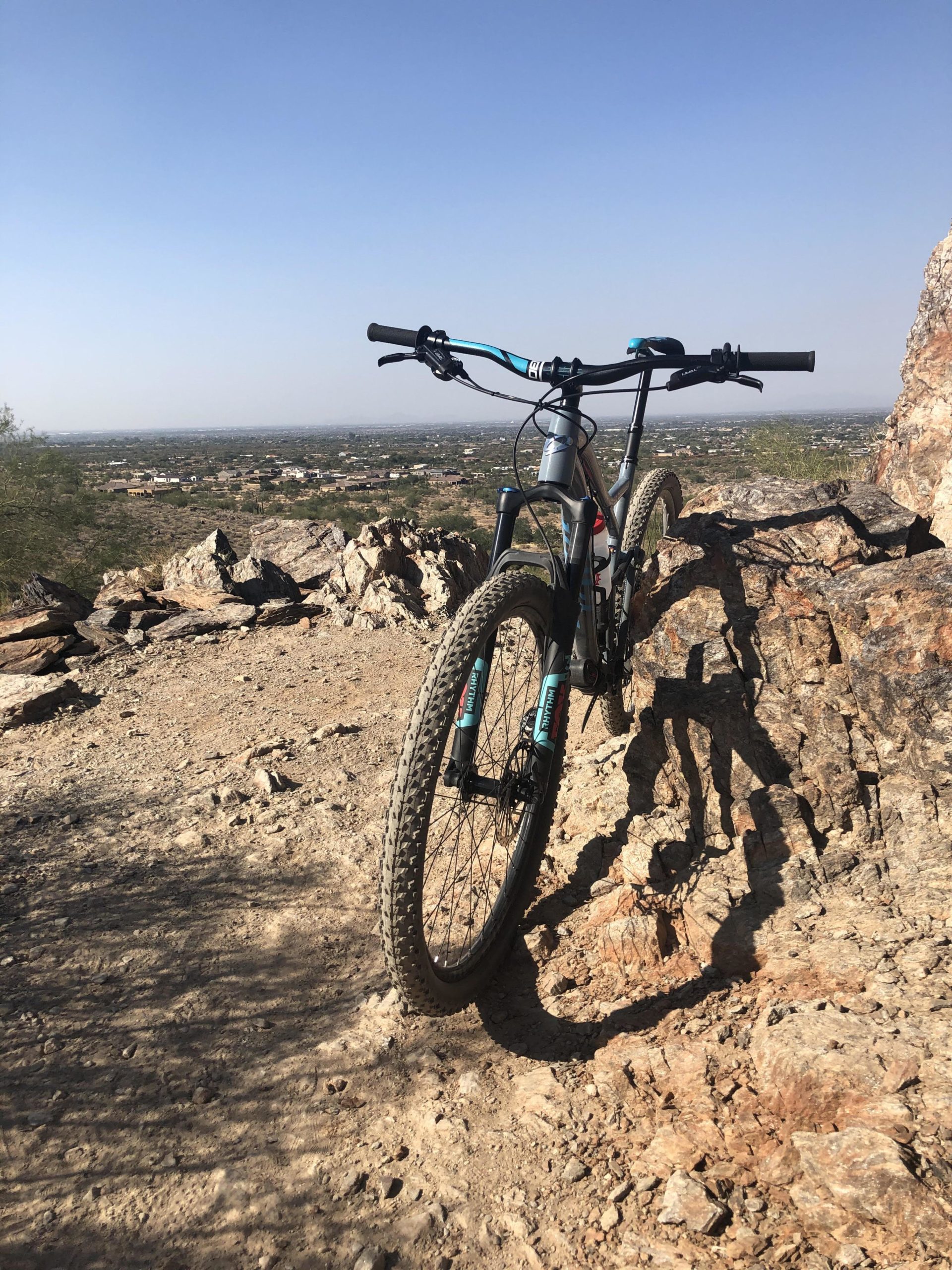 A mountain bike resting on rocky terrain, overlooking a vast landscape with distant buildings and hills under a clear blue sky. San Tan mountain bike trail.