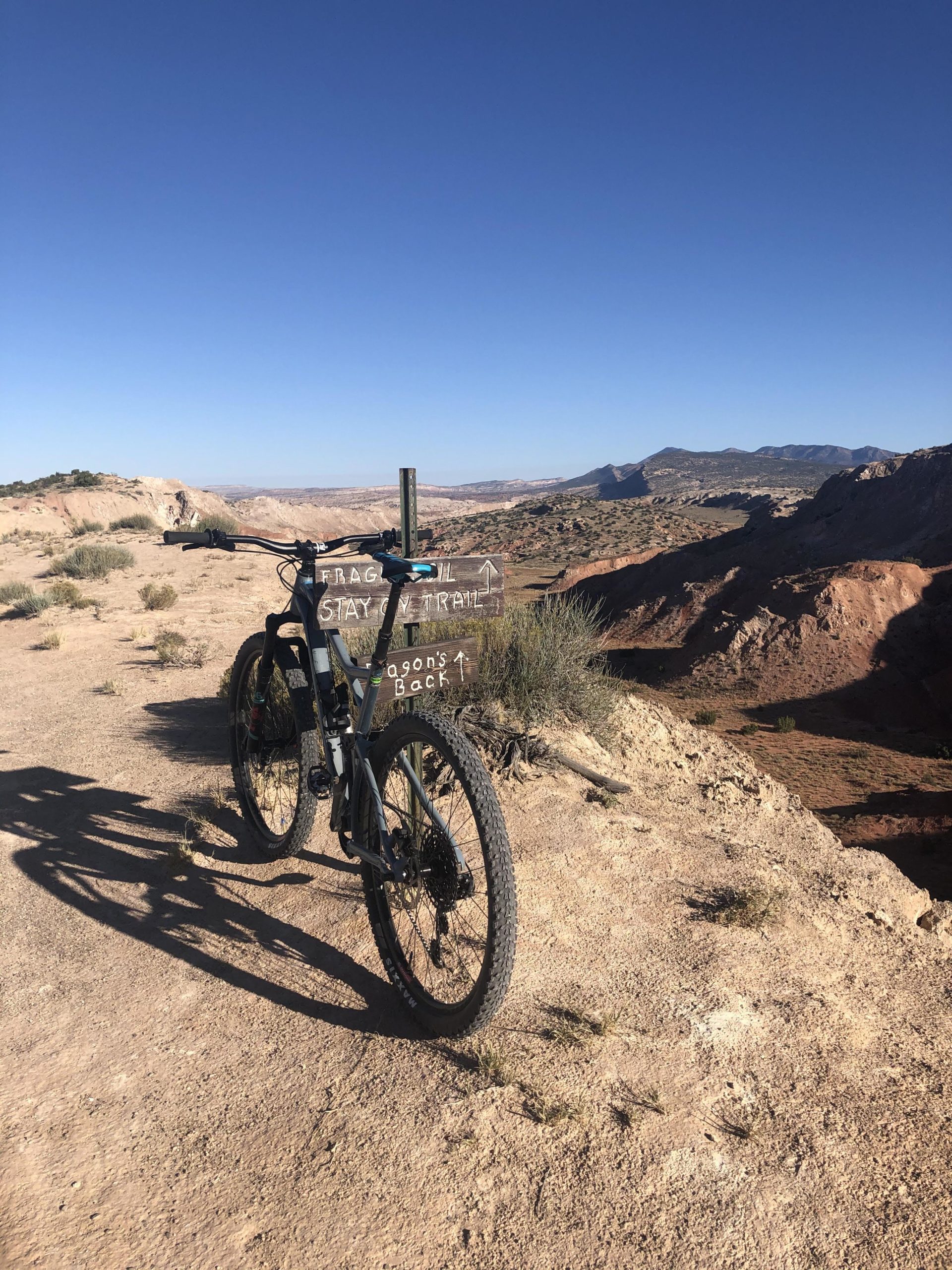 A mountain bike parked on a dirt trail with a sign indicating the direction to "Dragon's Back." The background features a vast landscape of rolling hills and rocky formations under a clear blue sky. Scrub vegetation is present along the trail. White Ridge Bike Trails mountain bike trail.