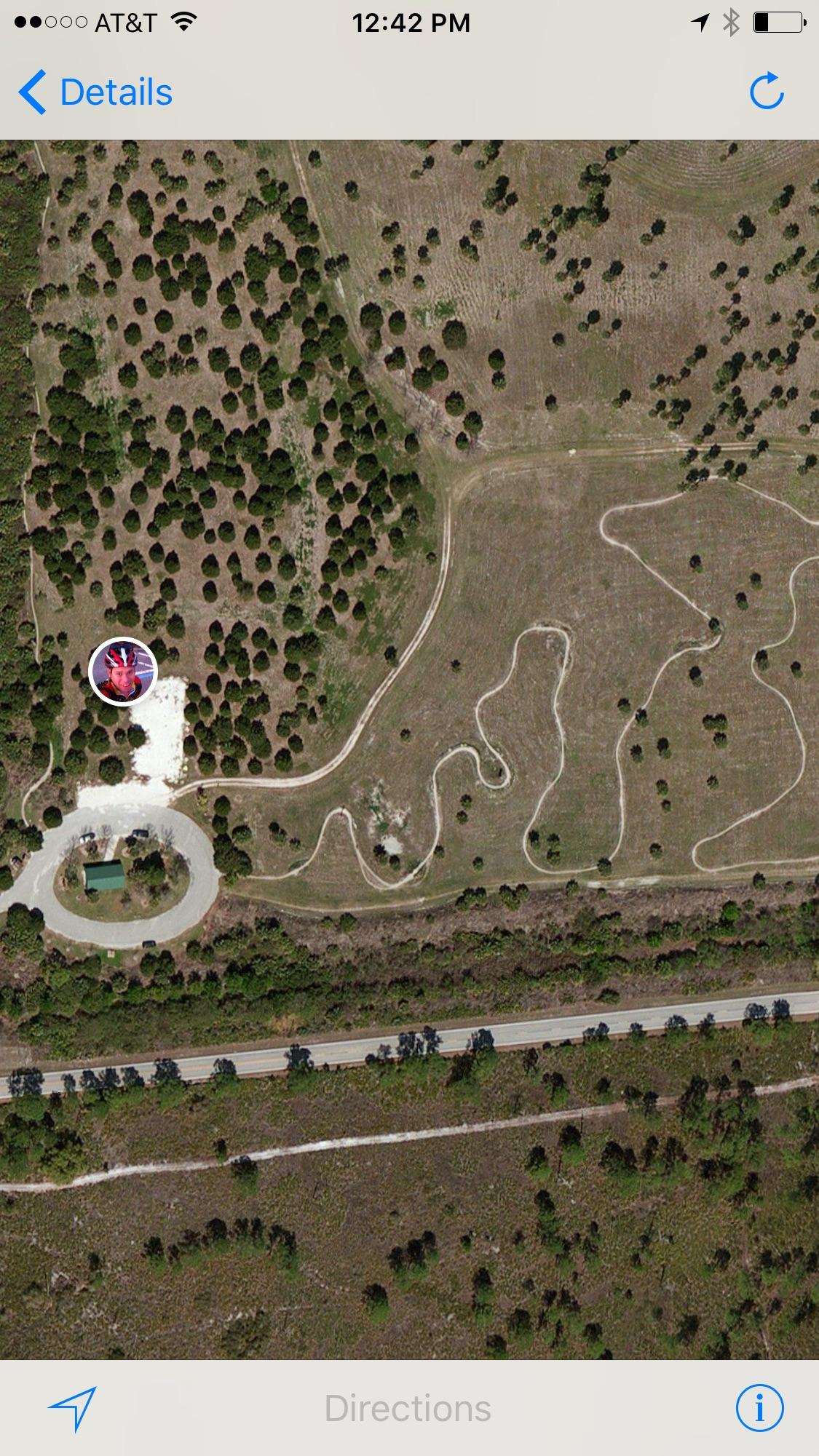 Aerial view of a rural area featuring winding dirt paths through scattered trees, with a circular road and a building at the bottom left. A circular icon is overlaid on the image, suggesting a specific location or point of interest.