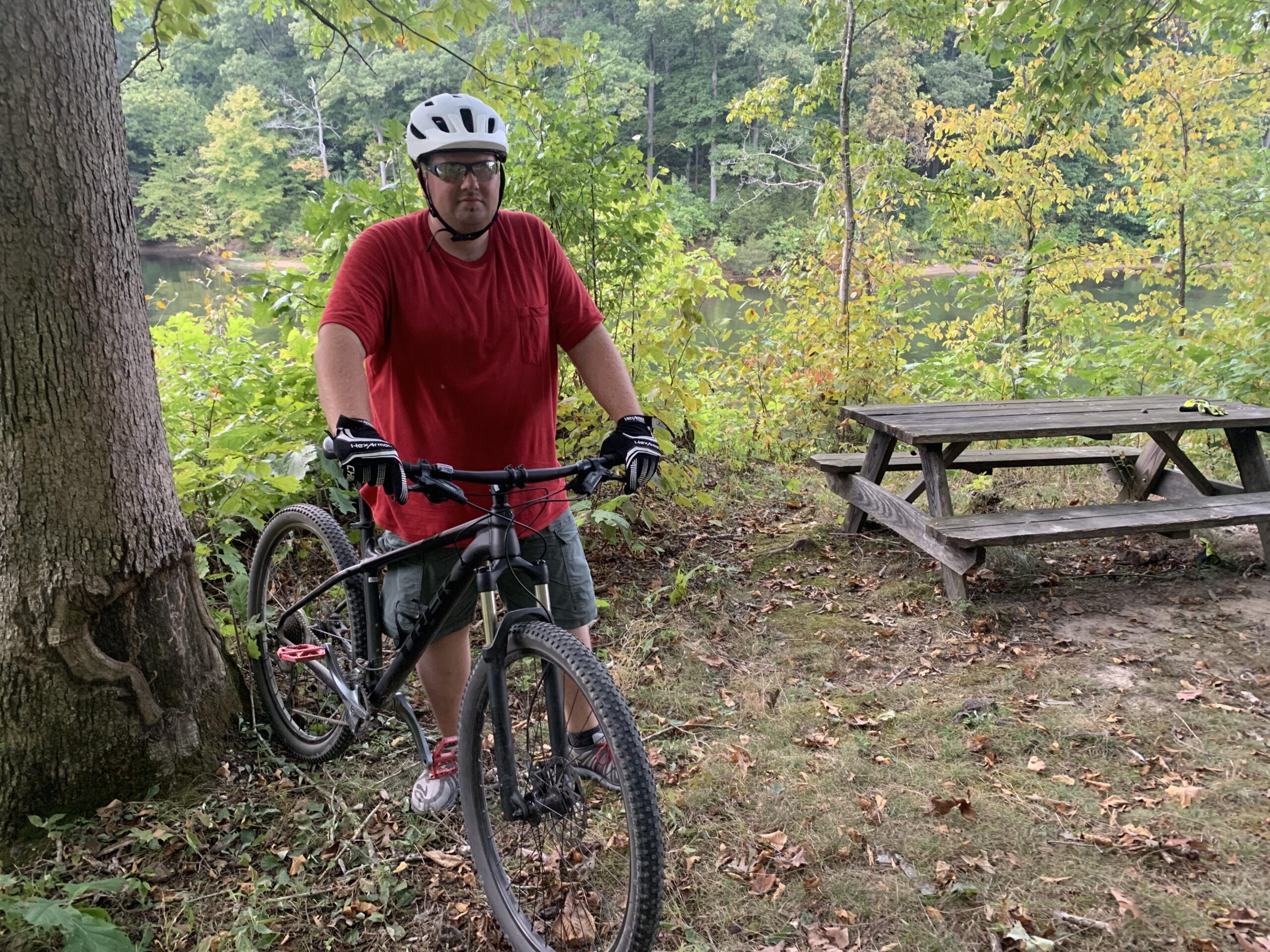 Trek X Caliber 7: A person wearing a helmet and sunglasses stands next to a mountain bike, leaning against a tree in a scenic outdoor area. They are dressed in a red shirt and shorts, surrounded by greenery and a picnic table in the background. A calm body of water is visible beyond the foliage.