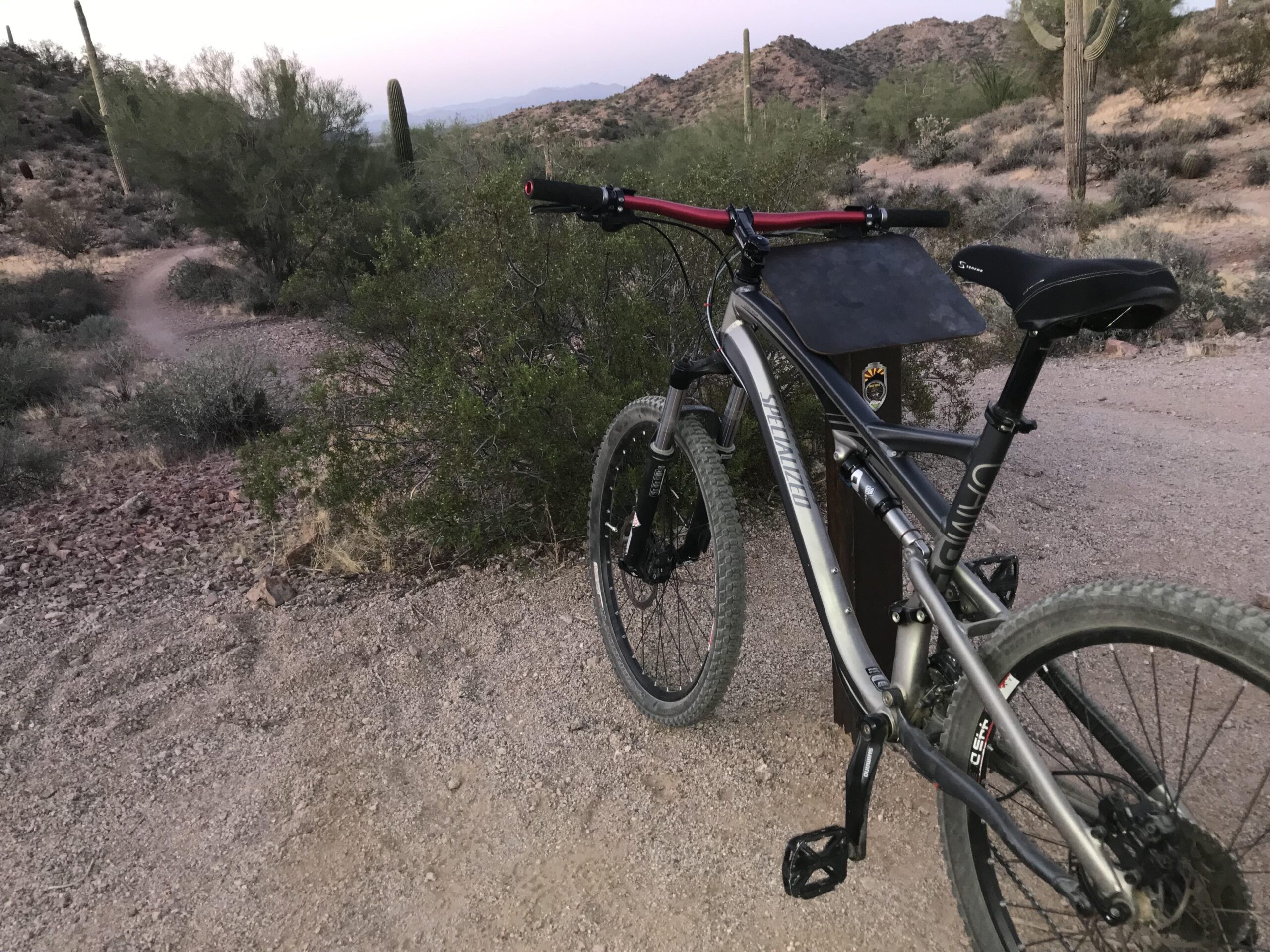 Specialized Camber: A mountain bike parked on a dirt trail in a desert landscape, surrounded by shrubs and cacti, with rugged hills in the background during twilight.