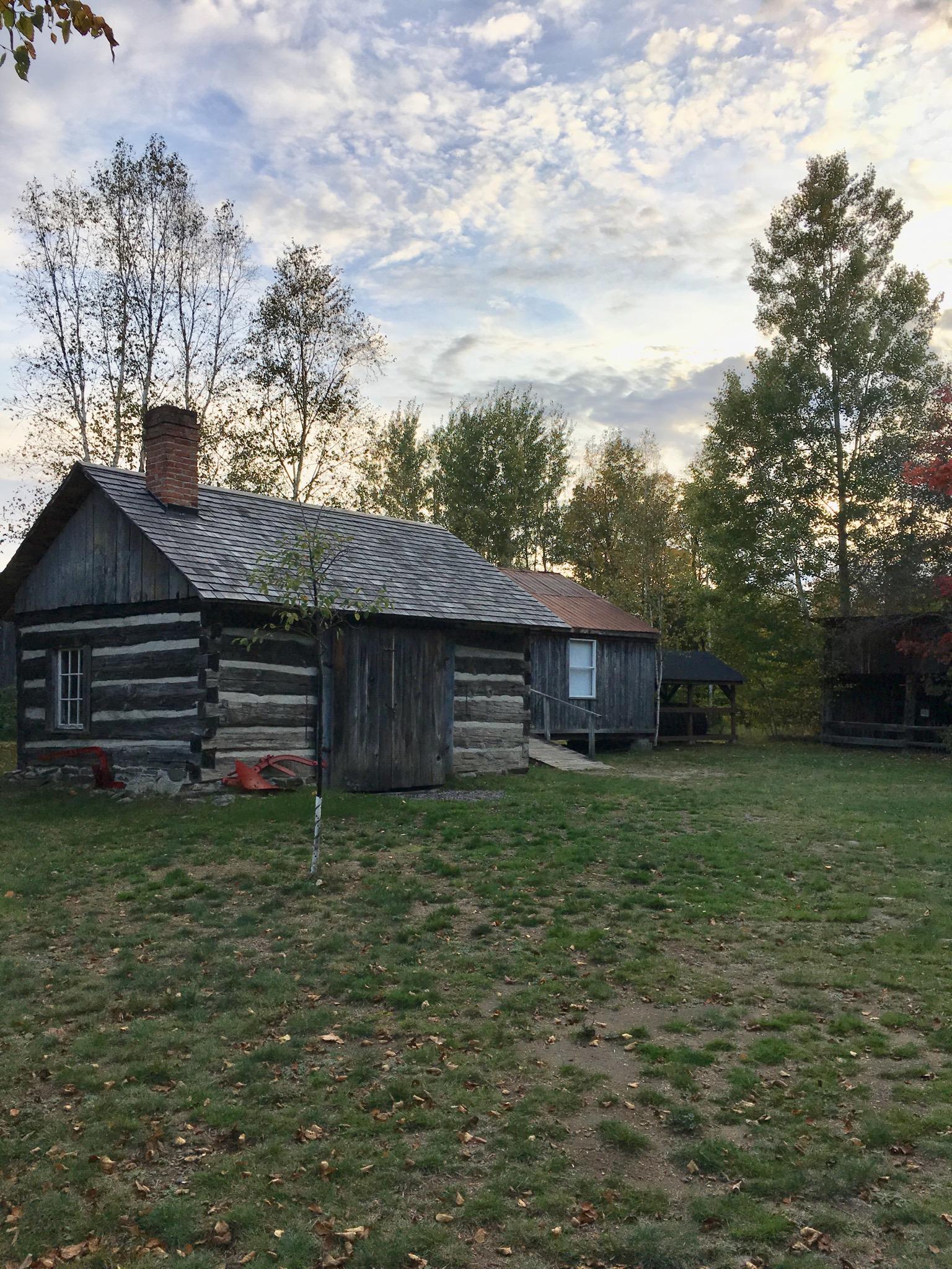 A rustic wooden log cabin surrounded by tall trees and a grassy area, set against a cloudy sky. The cabin features a sloped roof and a chimney, with a small porch area and another building visible in the background. Fallen leaves are scattered on the ground, suggesting autumn season. Glebe Park mountain bike trail.