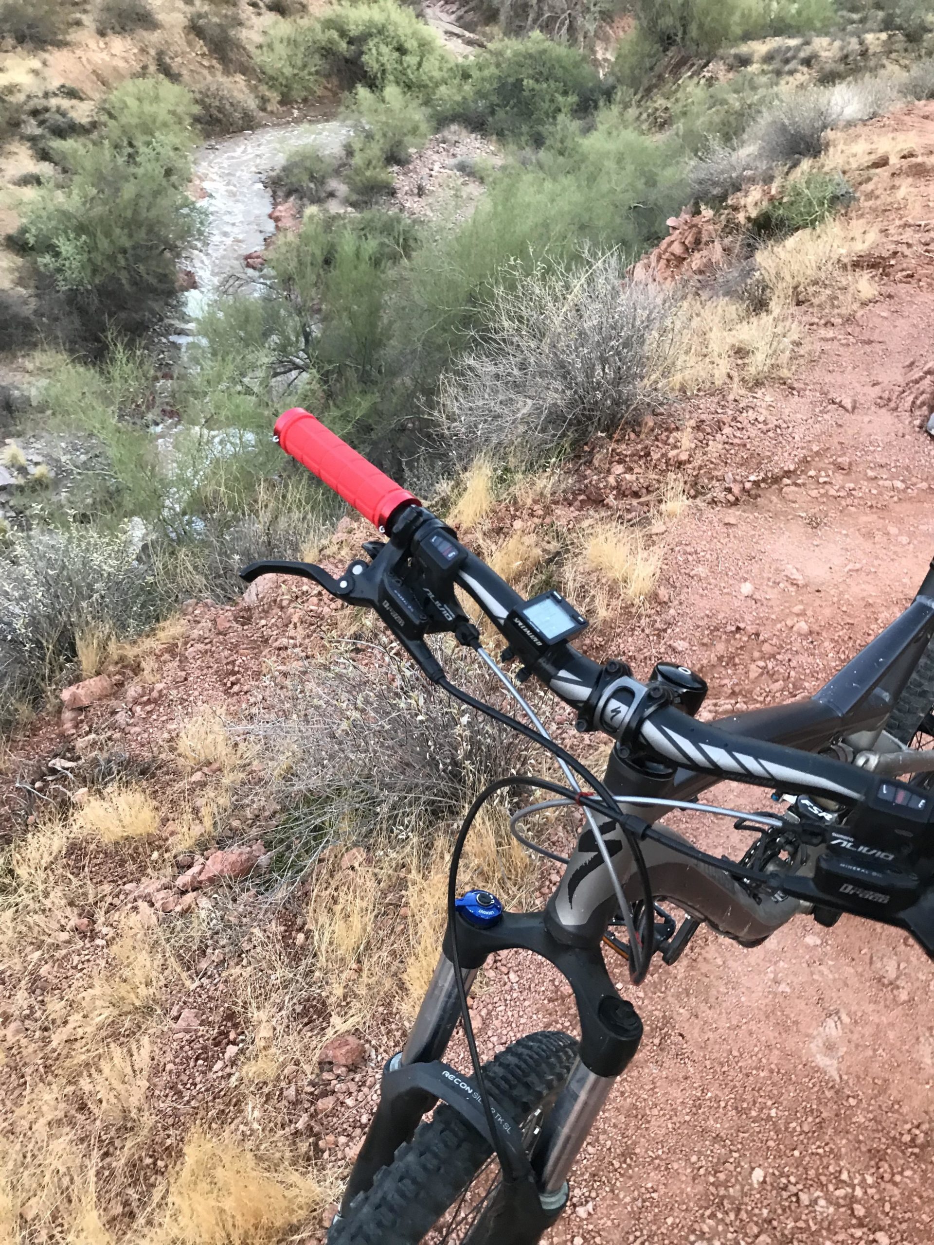 Close-up view of a mountain bike handlebar with a red grip, showing the brake lever, gear shifter, and display screen. In the background, a winding stream can be seen surrounded by green shrubs and rocky terrain. The ground is mostly dry and reddish-brown with patches of grass. Hawes Loop mountain bike trail.
