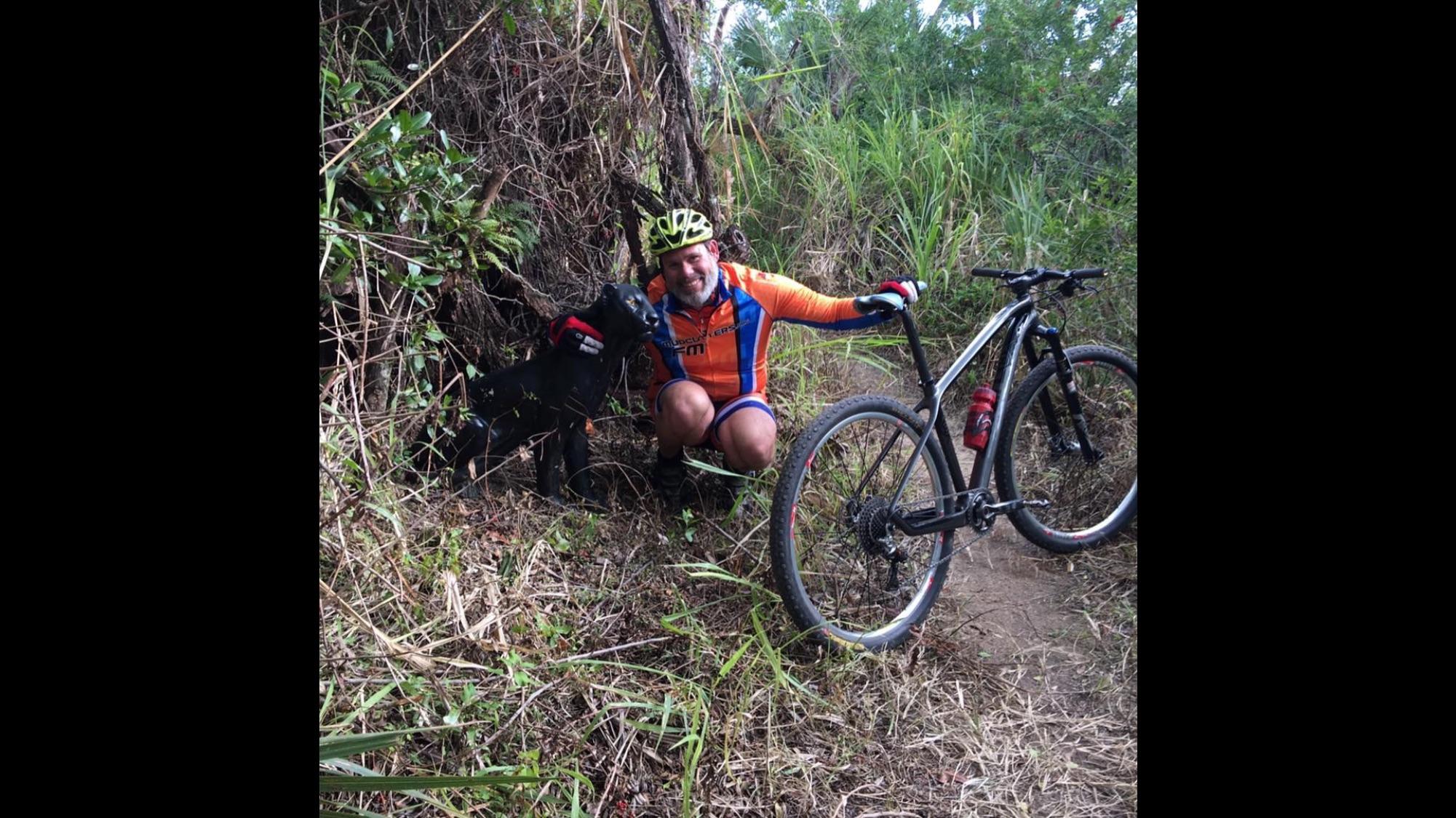 A smiling person in a colorful cycling outfit kneels next to a black dog in a forested area, with a mountain bike resting nearby. Lush greenery and foliage surround them, creating a natural outdoor setting.