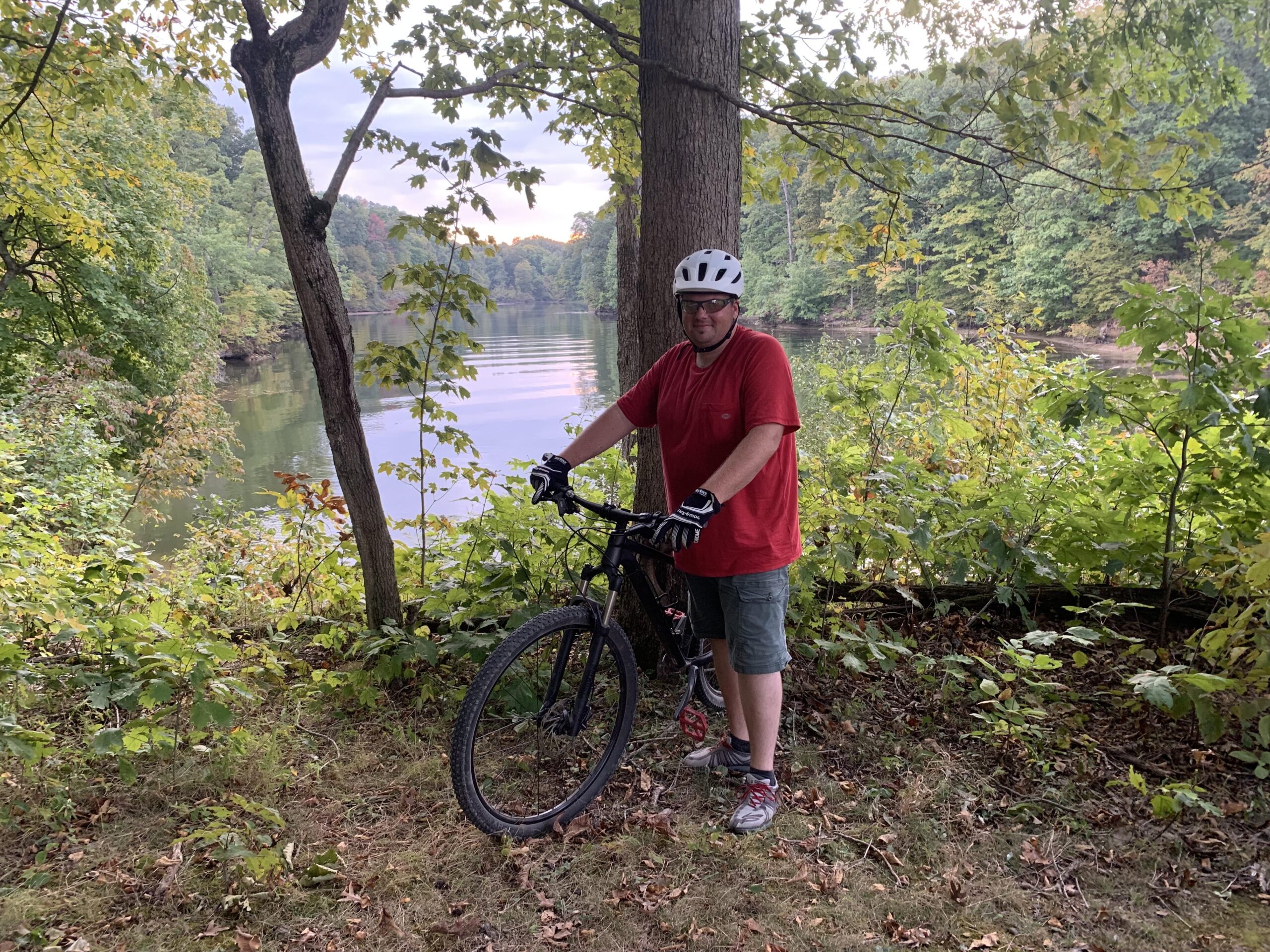 Trek X Caliber 7: A person wearing a red shirt, shorts, and a white helmet poses next to a mountain bike in a lush, green outdoor setting. In the background, a calm body of water reflects the surrounding trees, creating a serene atmosphere. The scene showcases the beauty of nature with colorful foliage typical of autumn.