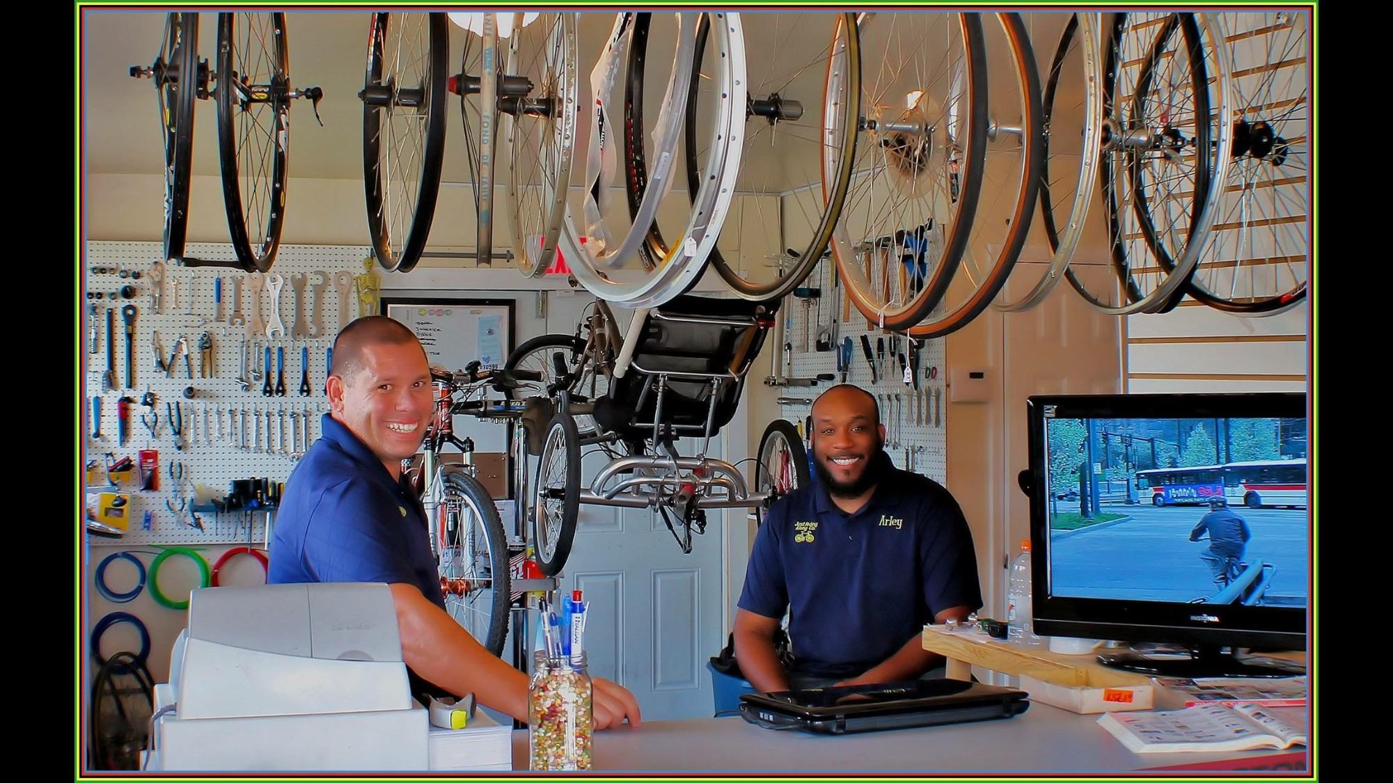 Two smiling employees are sitting at a counter in a bicycle shop, surrounded by bike wheels hanging from the ceiling and a variety of tools displayed on the wall behind them. One employee is wearing a navy blue shirt with "Arley" embroidered on it, while the other is in a similar shirt. A television screen is visible, showing a street scene with a cyclist in the background. The shop has a welcoming atmosphere with colorful decorations and organized tools.
