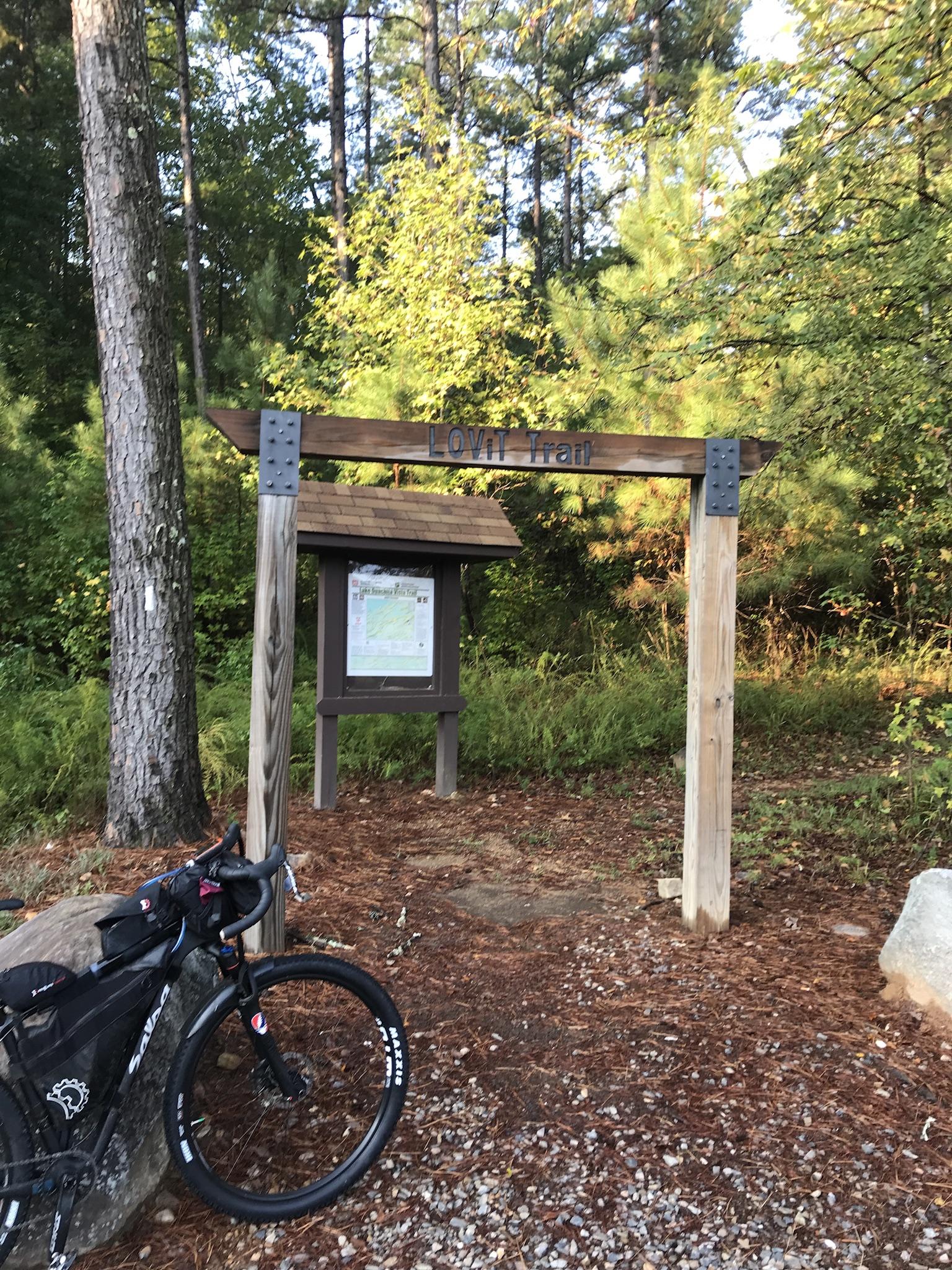 Wooden trailhead sign reading "LOVI Trail" with a trail map displayed nearby, surrounded by lush greenery and tall trees. A bicycle is parked beside a large rock in the foreground on a gravel path. Lake Ouachita Vista Trail (LOViT) mountain bike trail.