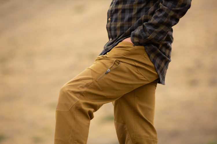 A person wearing a plaid shirt and tan outdoor pants, standing with one leg bent and their hand in their pocket, against a blurred outdoor background.