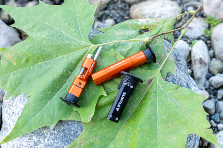Two outdoor tools are positioned on a large green maple leaf, with a background of rocks and pebbles. The tools include one orange and black device, and another predominantly orange tool, both designed for camping or hiking use. Natural elements in the scene enhance the outdoor setting.