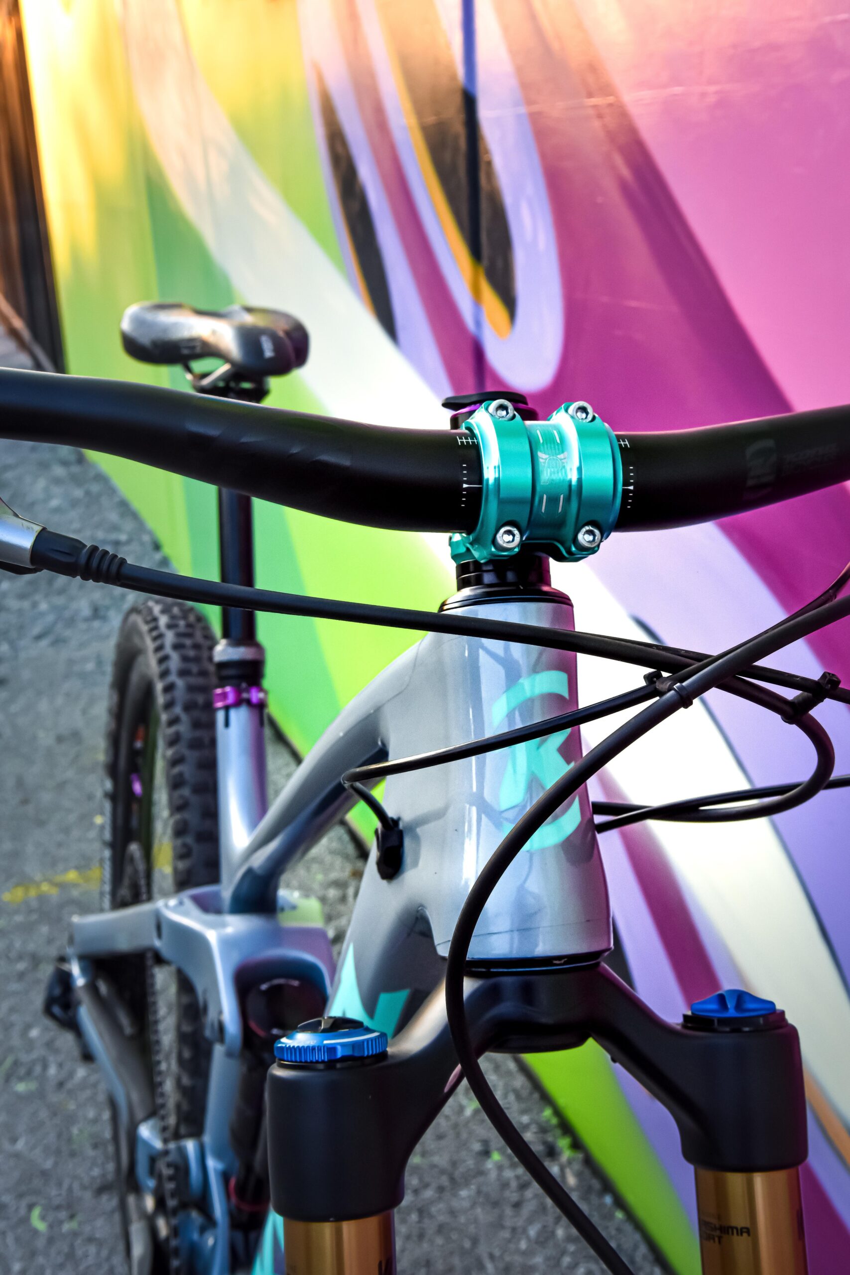 Kona Process 153: A close-up view of a mountain bike's handlebars and stem, featuring a turquoise stem clamp and intricate cable routing. The bike is positioned against a colorful, abstract mural with vibrant green, pink, and purple hues.