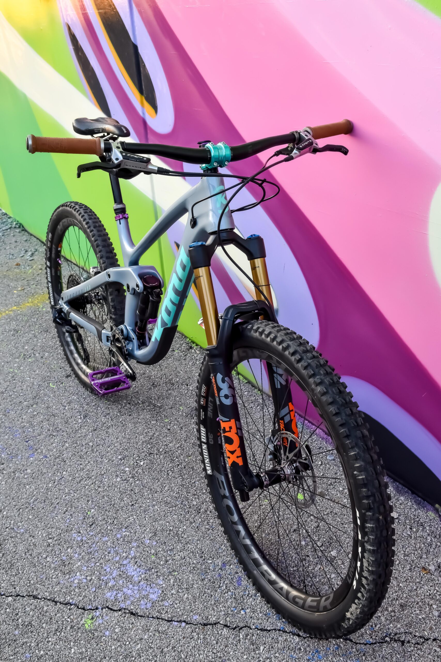 Kona Process 153: A close-up image of a mountain bike with a grey frame, featuring gold front suspension forks and purple pedals. The handlebars have brown grips, and the bike is positioned against a vibrant, colorful mural with green and pink elements in the background. The ground is paved with textured asphalt.