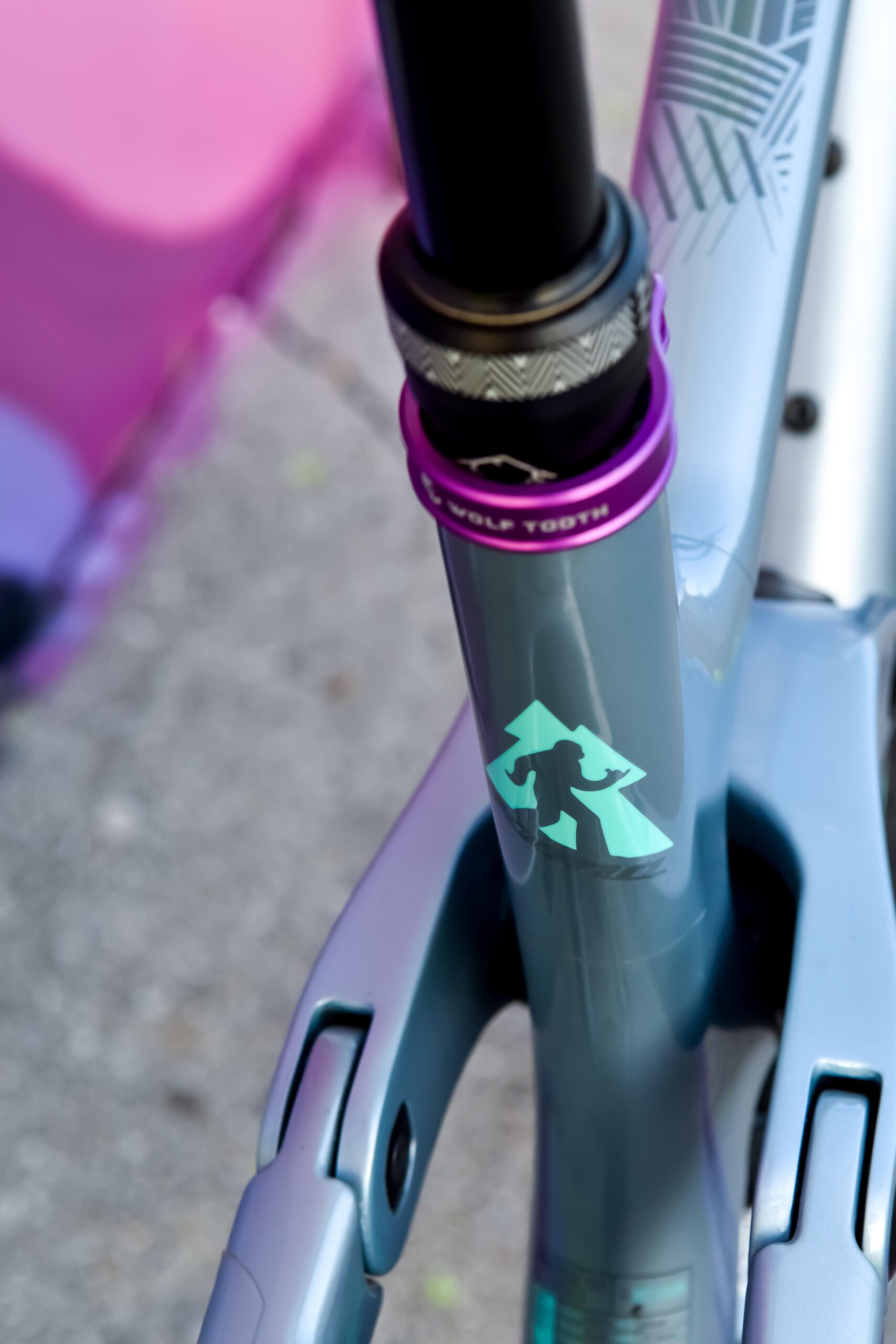 Kona Process 153: Close-up of a bike frame featuring a teal design element of a mountain silhouette with a playful character. The stem showcases a purple headset cap labeled "Wolf Tooth," and the bike has a modern, sleek design with a gradient effect in shades of gray and purple. The background is slightly blurred, highlighting the frame details.