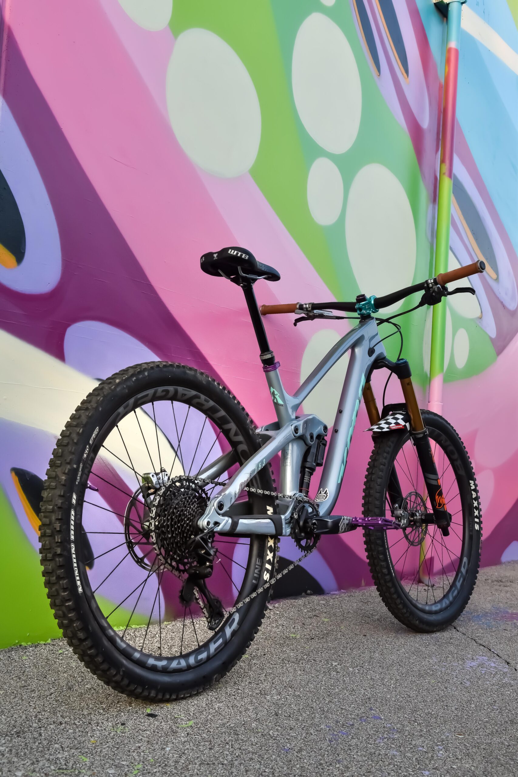 Kona Process 153: A mountain bike with a silver and blue frame is positioned at an angle against a vibrant, multi-colored graffiti wall featuring pink and green abstract patterns. The bike has prominent black tires with intricate tread patterns and a comfortable saddle. The scene combines outdoor sport with urban street art.