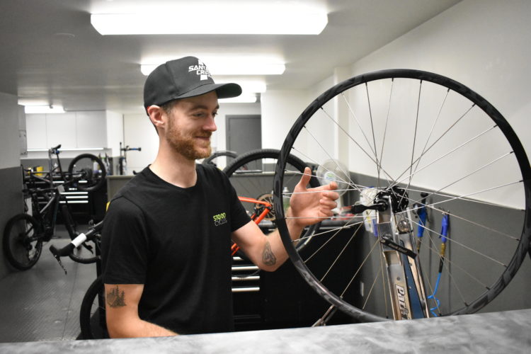 A smiling mechanic holding a bicycle wheel in a bike shop, with various bicycles and tools visible in the background.