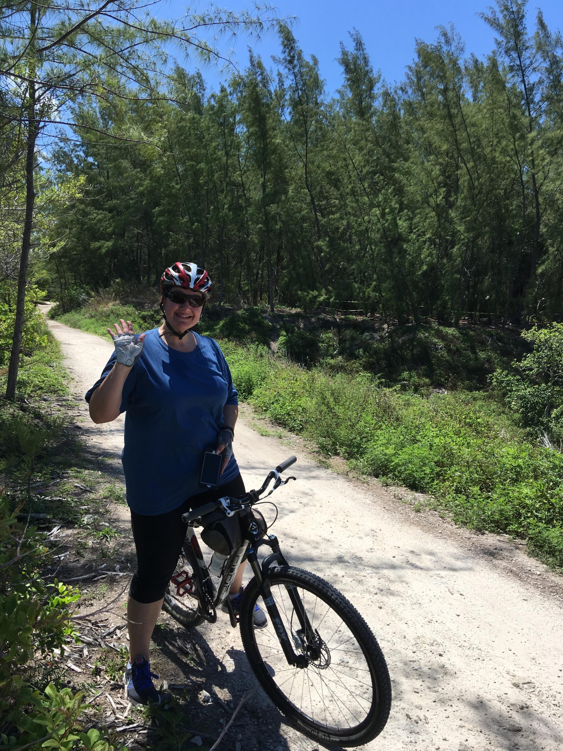 A smiling person wearing a helmet and sunglasses is standing next to a mountain bike on a dirt path surrounded by greenery and tall trees. They are waving with one hand and holding a smartphone in the other under a clear blue sky.