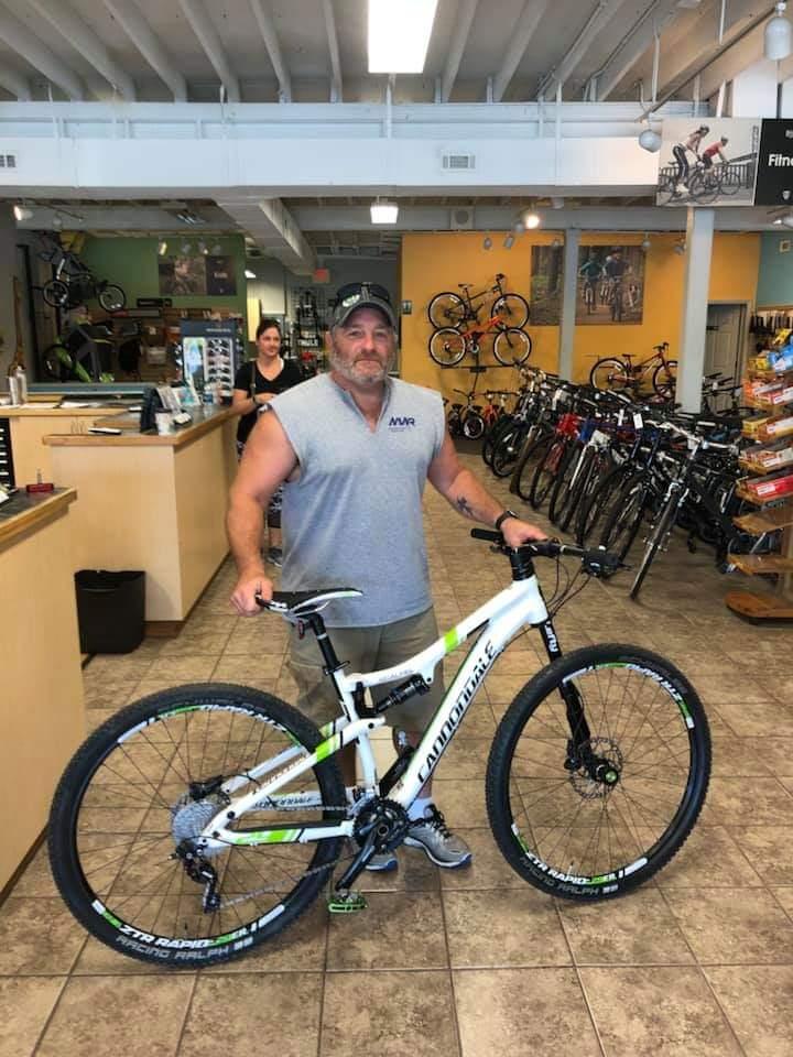 Cannondale Scalpel 29er 3: A man stands in a bike shop holding a white mountain bike with green and black accents. He is wearing a gray sleeveless shirt and gray shorts. The store has a variety of bicycles displayed in the background, and there is a sales counter to the left with another person nearby. The setting is bright and welcoming, with a tiled floor.