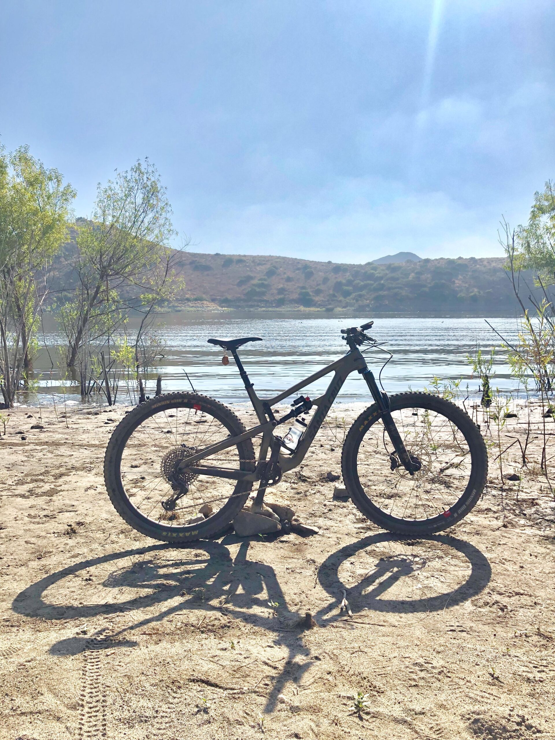 Santa Cruz Hightower LT: A mountain bike parked on sandy ground near a calm lake, with hills and trees in the background under a clear blue sky. The bike casts a long shadow on the sand.