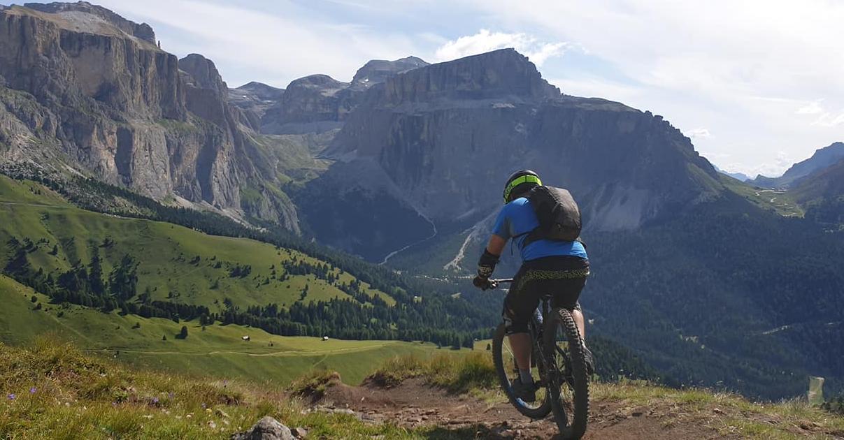 Rocky Mountain Pipeline: A mountain biker rides along a rugged trail with vast green hills and towering mountains in the background under a partly cloudy sky.