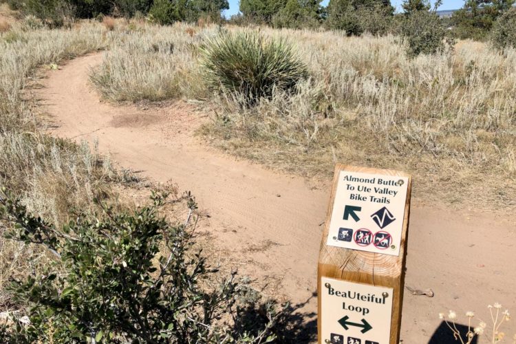 A wooden trail sign in a natural setting, indicating the directions to "Almond Butter to Ute Valley Bike Trails" and "BeaUteiful Loop." The sign features symbols for hiking and biking, with paths winding through grassy terrain and shrubs.