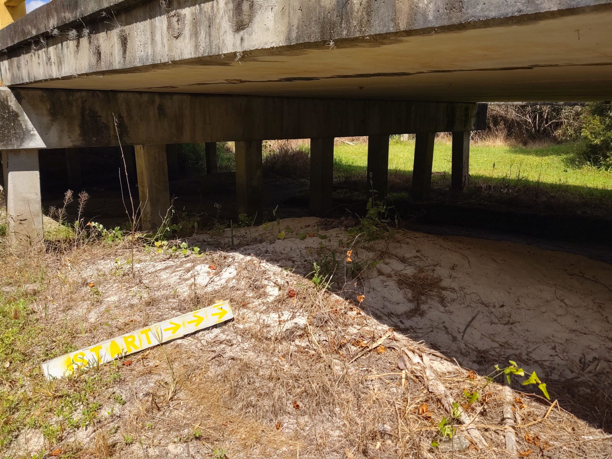 A sandy area beneath a concrete bridge, with a yellow sign reading "START" and arrows indicating direction. The scene is surrounded by overgrown grass and vegetation, with sunlight filtering through the structure above. North Port Mountain Bike Trails mountain bike trail.