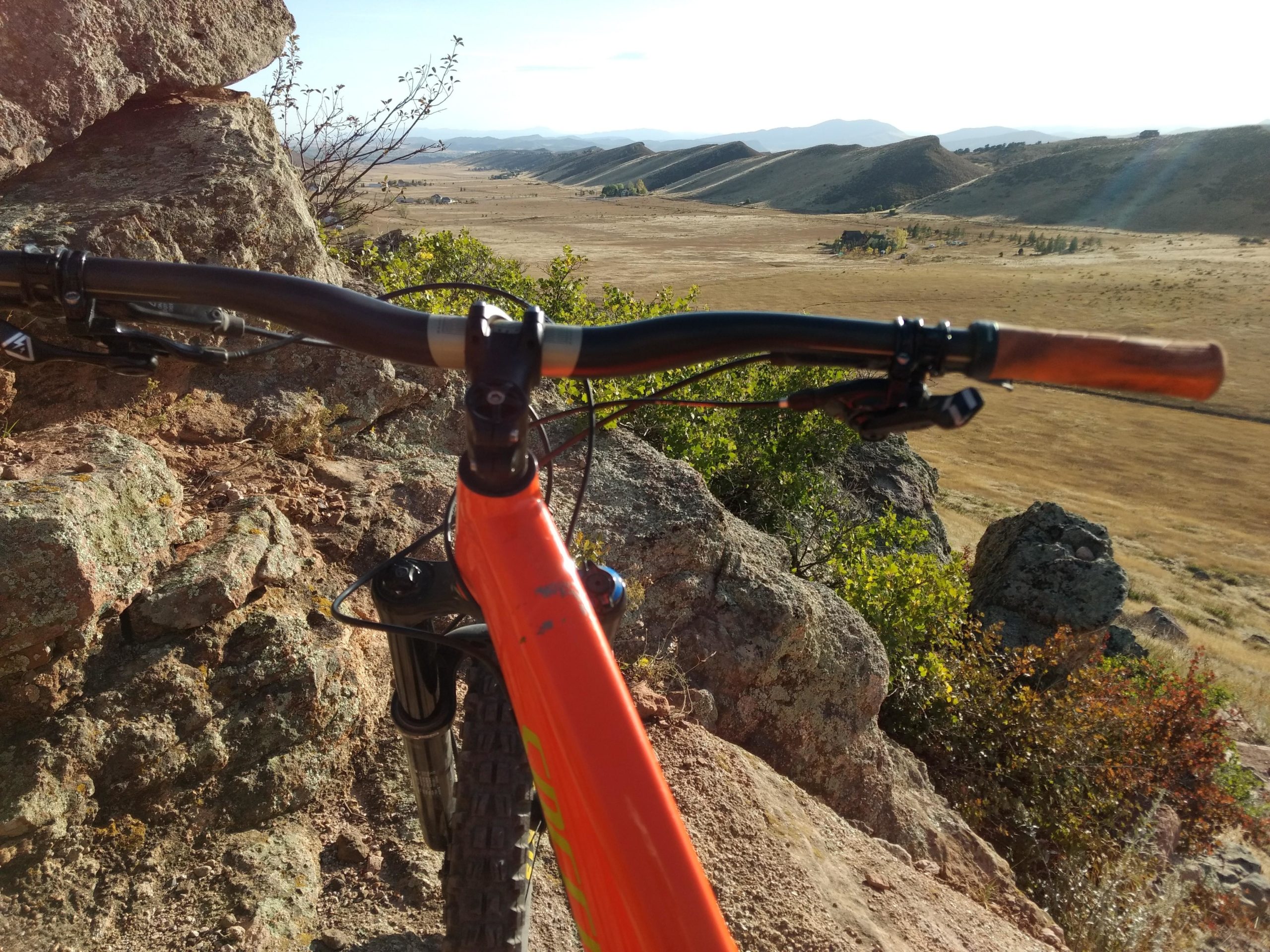 A close-up view of a mountain bike's handlebars, set against a scenic landscape of rolling hills and a grassy field in the background. The bike's bright orange frame is visible, along with rocky terrain and vegetation surrounding it. The sky is clear with a soft natural light illuminating the scene. Coyote Ridge mountain bike trail.