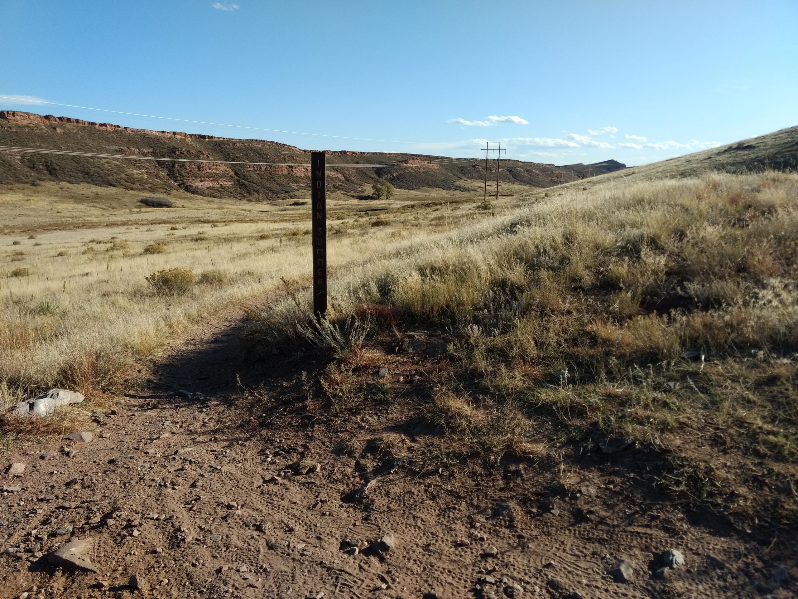 A grassy landscape with rolling hills under a clear blue sky. In the foreground, a dirt path leads towards a wooden post labeled "Indian Summer." Power lines run along the horizon, and rocky outcrops are visible in the distance. The scene captures a serene, natural setting. Indian Summer mountain bike trail.