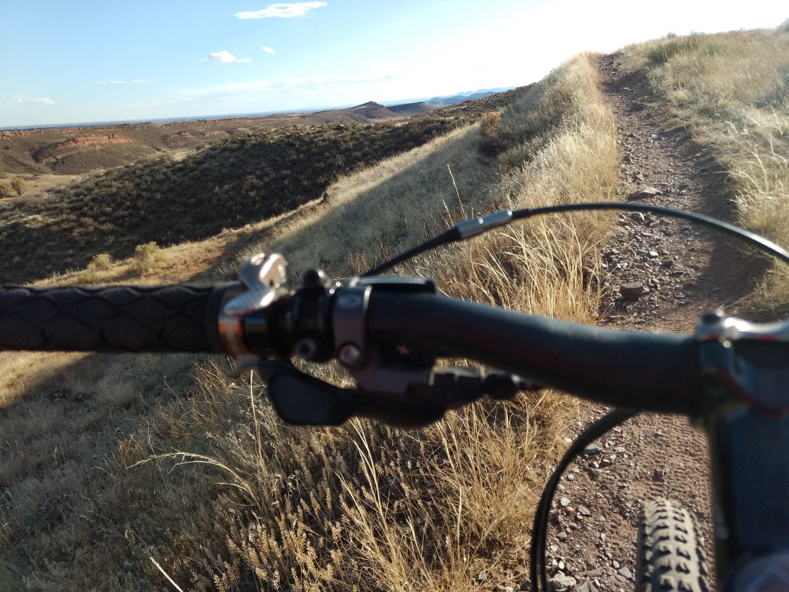 Mountain bike handlebar in the foreground with a gravel trail winding through dry, grassy terrain and hills in the background under a clear blue sky. Indian Summer mountain bike trail.