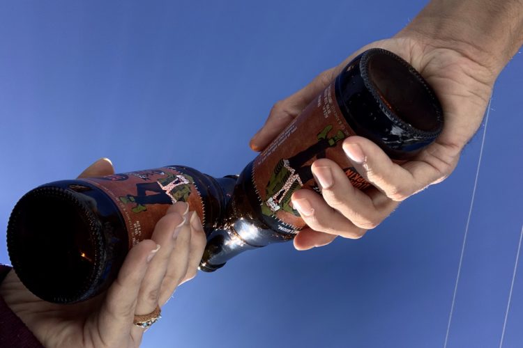 Two hands are holding brown glass beer bottles with colorful labels, set against a clear blue sky. One hand on the left has a ring and manicured nails, while the hand on the right has a more casual grip.