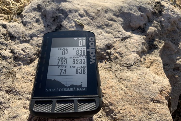 A close-up view of a Wahoo cycling computer resting on a rock, displaying metrics such as grade, elevation, temperature, and climbing statistics.