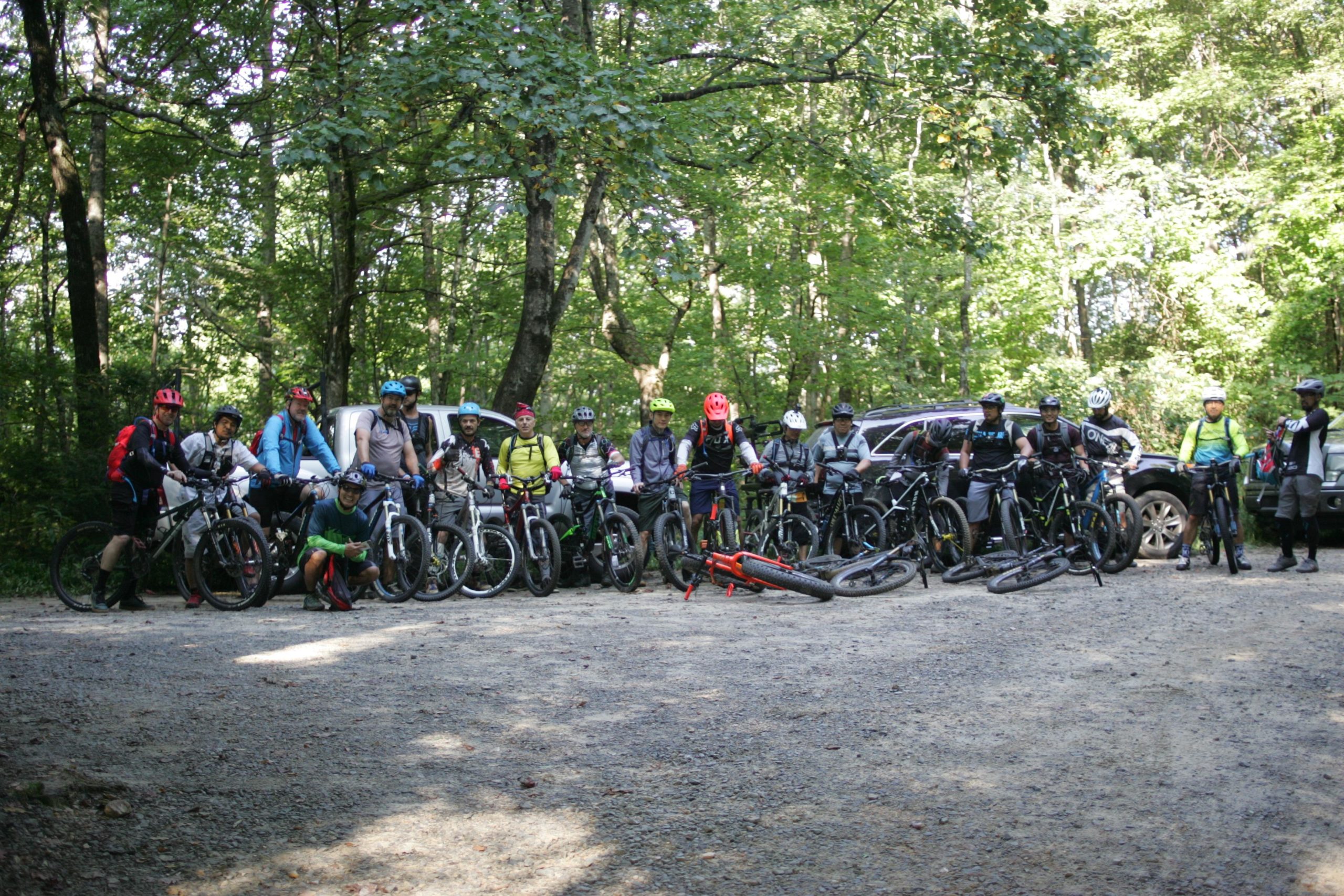 A group of mountain bikers gathered at a gravel parking area surrounded by lush green trees. The riders are posing with their bikes, wearing helmets and cycling gear, while a few vehicles are parked in the background. The scene captures the camaraderie and enthusiasm of the group as they prepare for a ride. Avery Creek / 327 mountain bike trail.