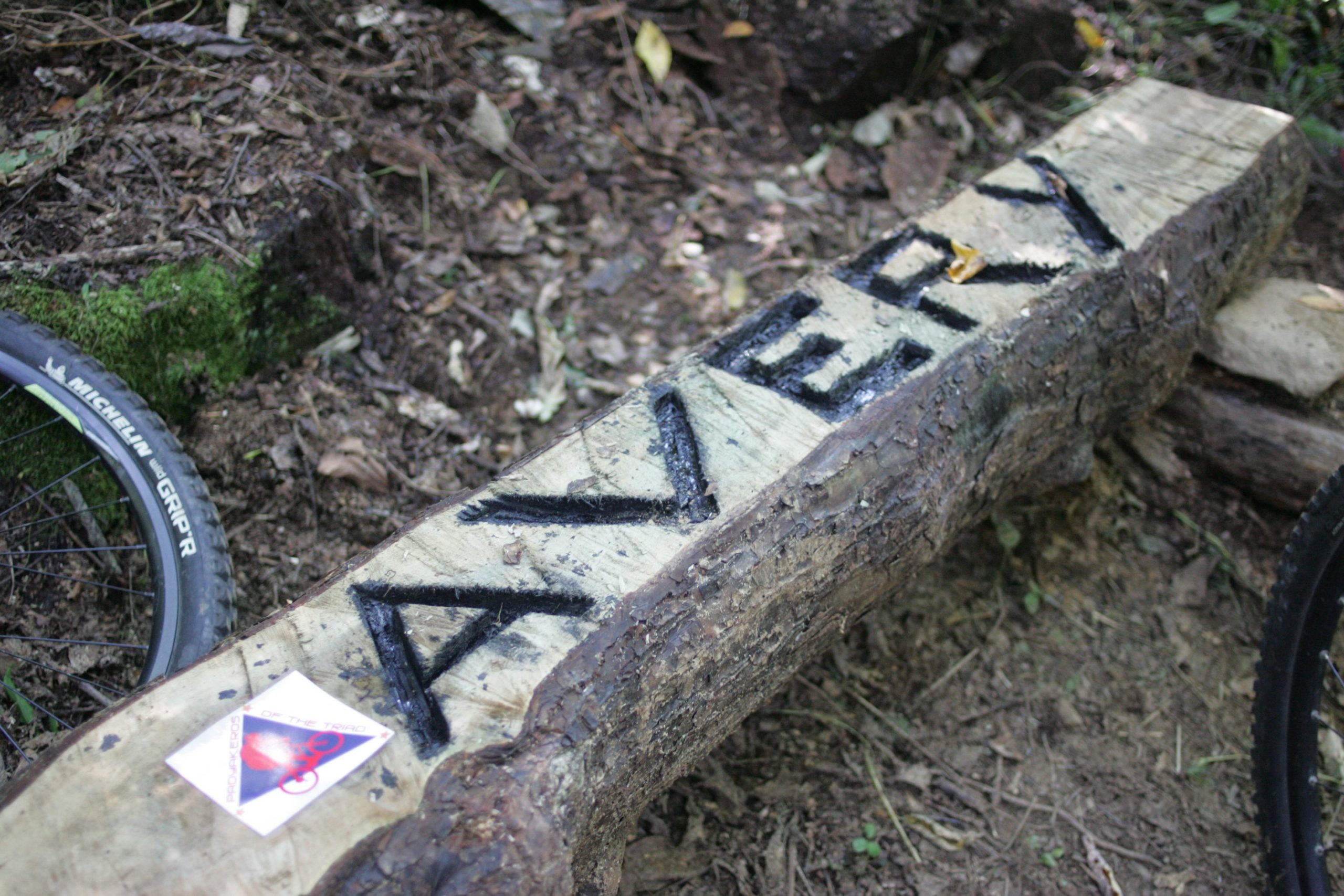 A wooden log carved with the name "Avery," placed on the ground in a forested area, surrounded by leaves and dirt. A mountain bike wheel is partially visible in the background, along with a sticker featuring a bike design. Avery Creek / 327 mountain bike trail.