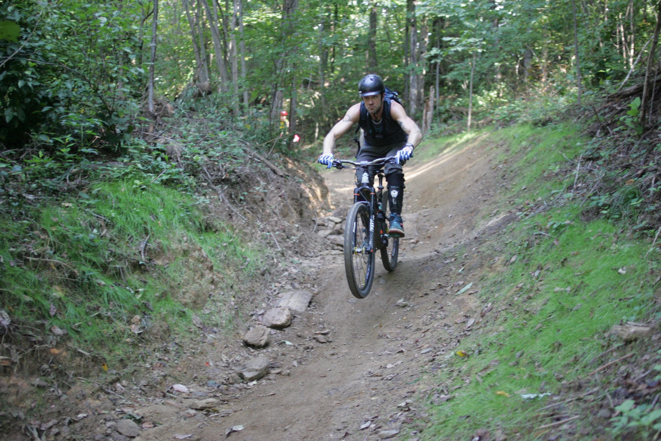 Mountain biker navigating a curved dirt trail in a forested area, surrounded by greenery and scattered rocks. The rider is wearing a helmet and protective gear, focused on maintaining balance while descending the path. Avery Creek / 327 mountain bike trail.