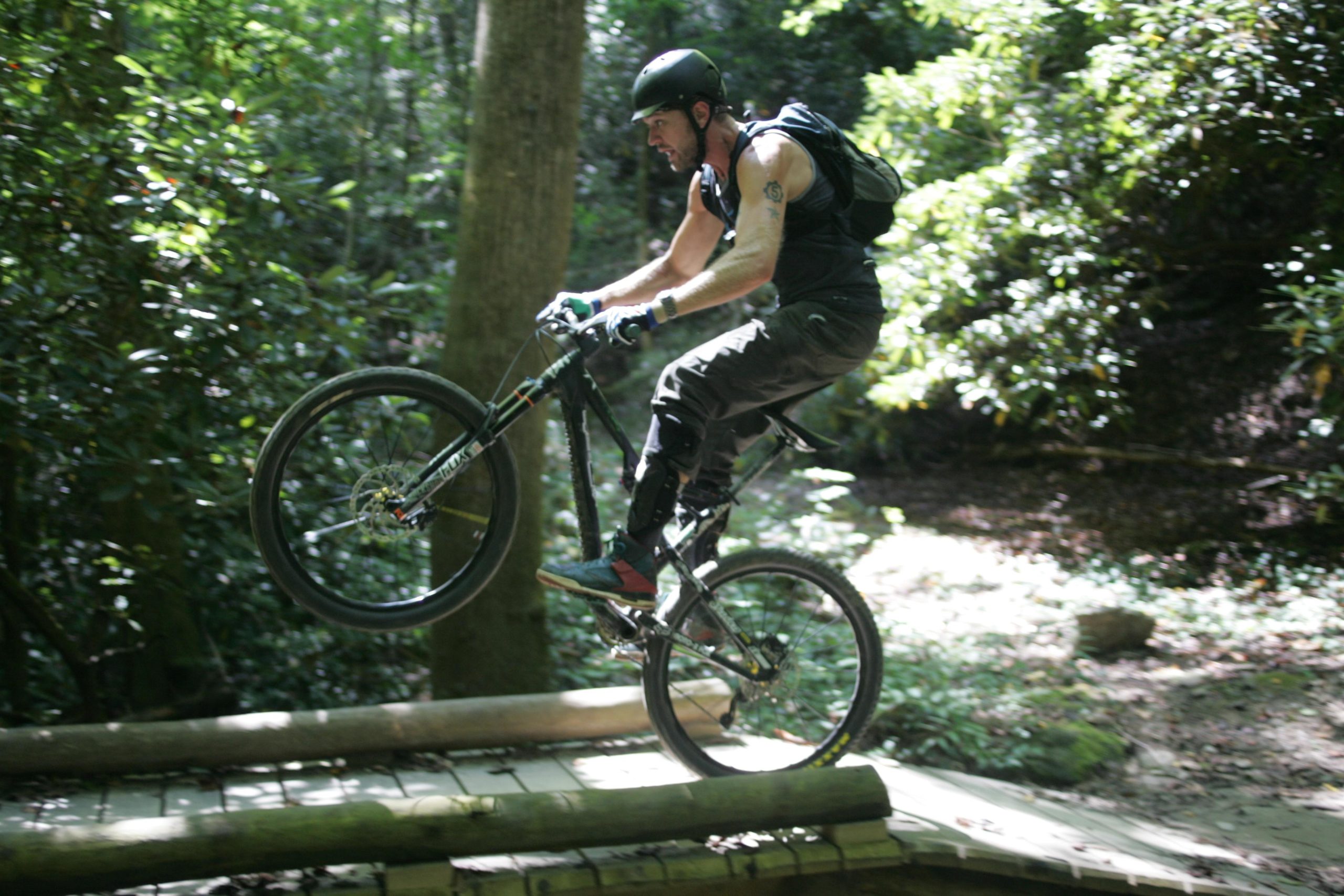 A mountain biker performing a wheelie on a wooden bridge in a lush, green forest. The cyclist is wearing a helmet and protective gear, and the background features trees and foliage, capturing an adventurous outdoor scene. Black Mountain mountain bike trail.