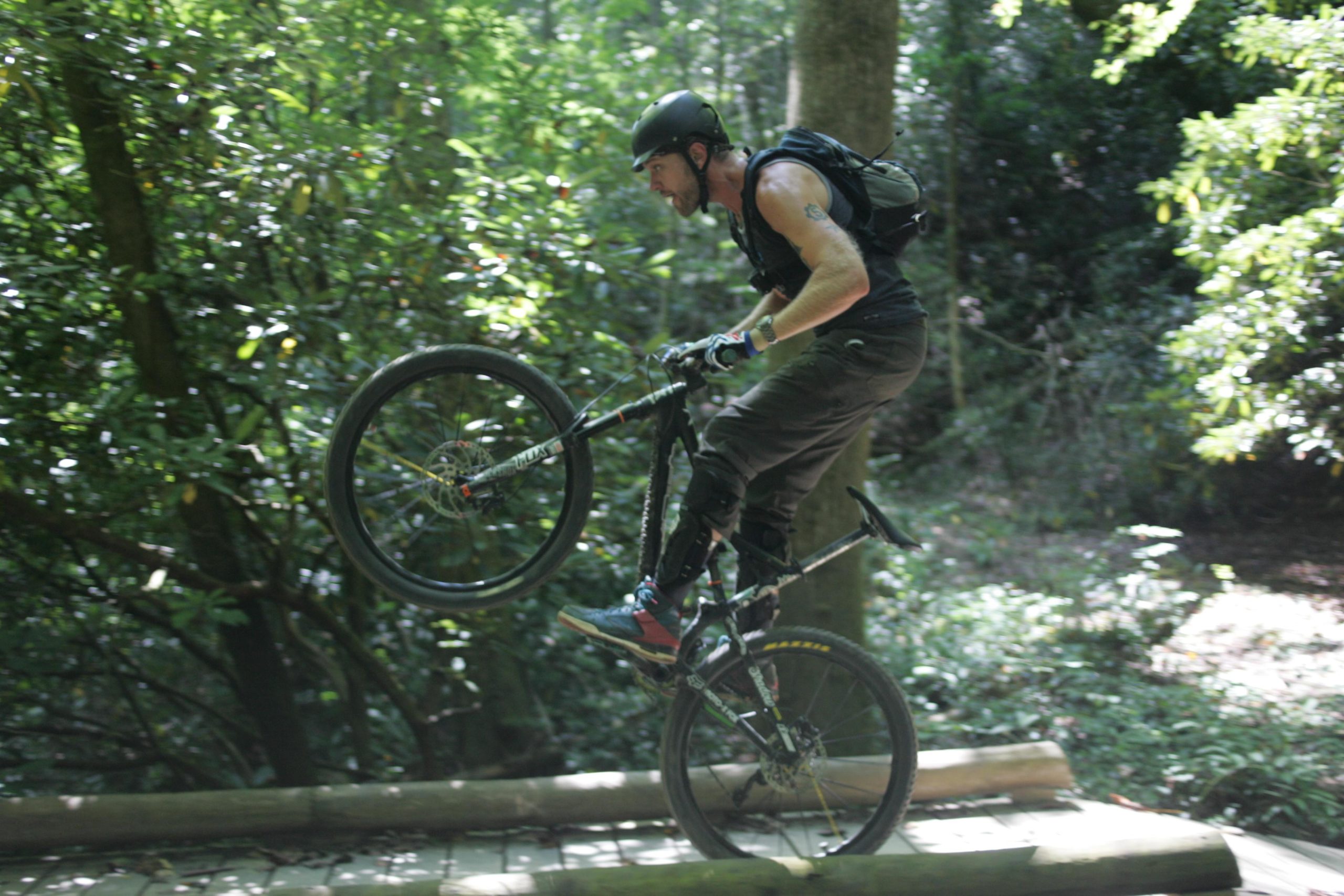 A mountain biker performing a stunt on a wooden trail in a lush forest setting, with one wheel lifted off the ground while balancing on the bike. The rider is wearing a helmet, gloves, and dark clothing, surrounded by greenery. Black Mountain mountain bike trail.