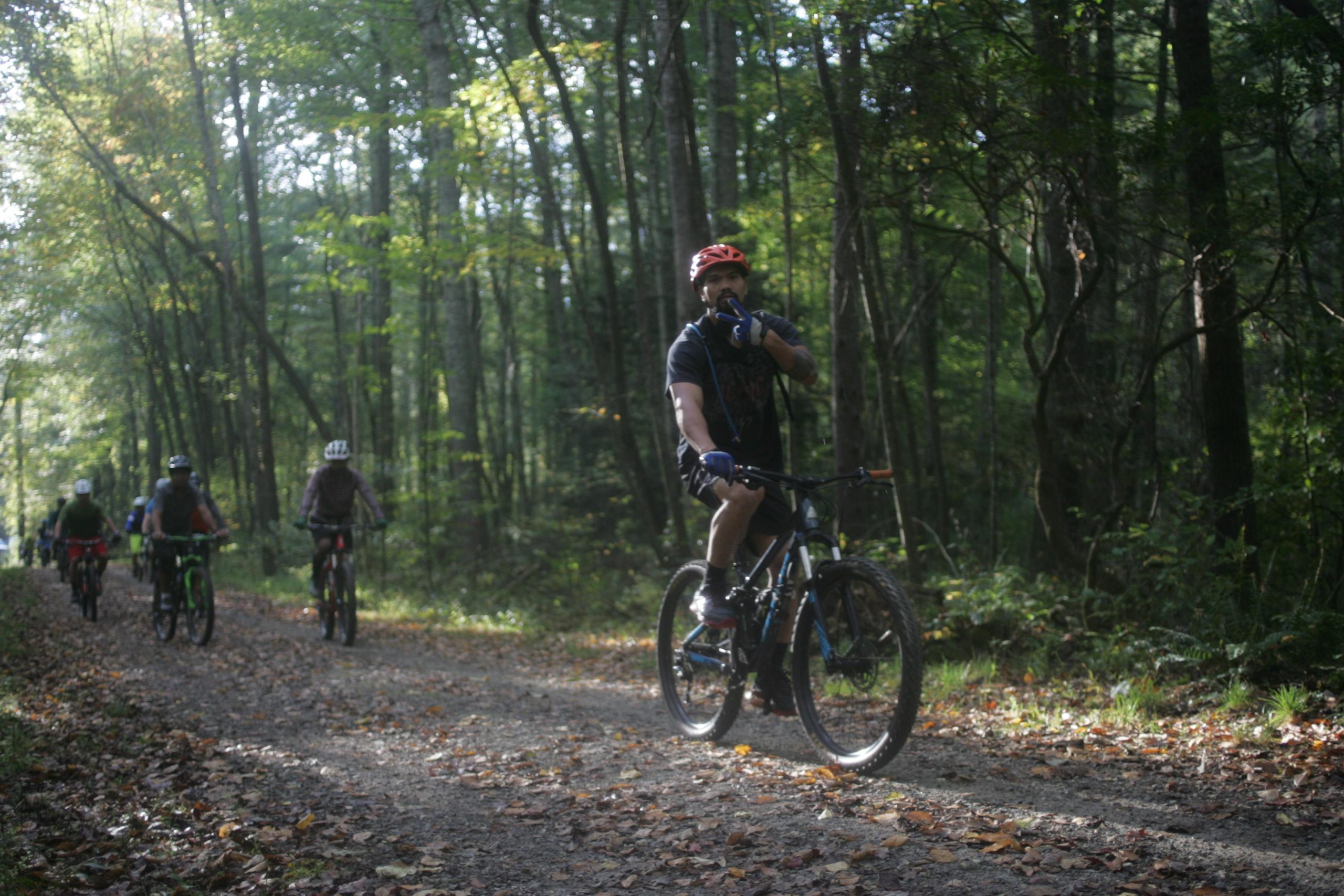 A group of mountain bikers riding along a gravel path in a forest setting. The foreground features a man on a bike, wearing a red helmet and gesturing with his finger to his lips, signaling for quiet. Surrounding trees are lush and green, with sunlight filtering through the leaves, creating a serene atmosphere. Bent Creek mountain bike trail.