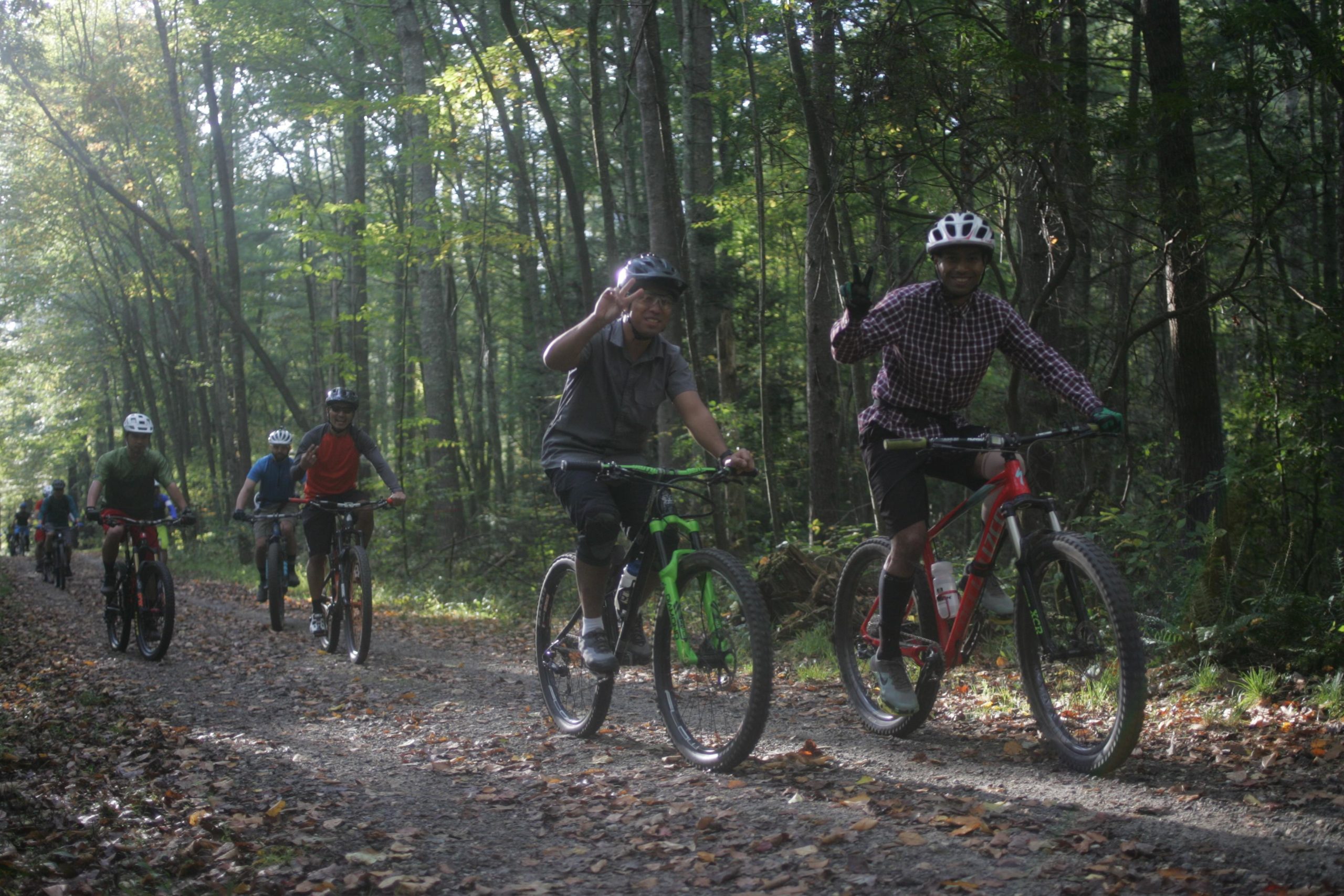 A group of mountain bikers riding on a dirt path through a wooded area, with two cyclists in the foreground smiling and giving a peace sign. The scene is illuminated by soft, natural light filtering through the trees, creating a vibrant, outdoor atmosphere. Bent Creek mountain bike trail.