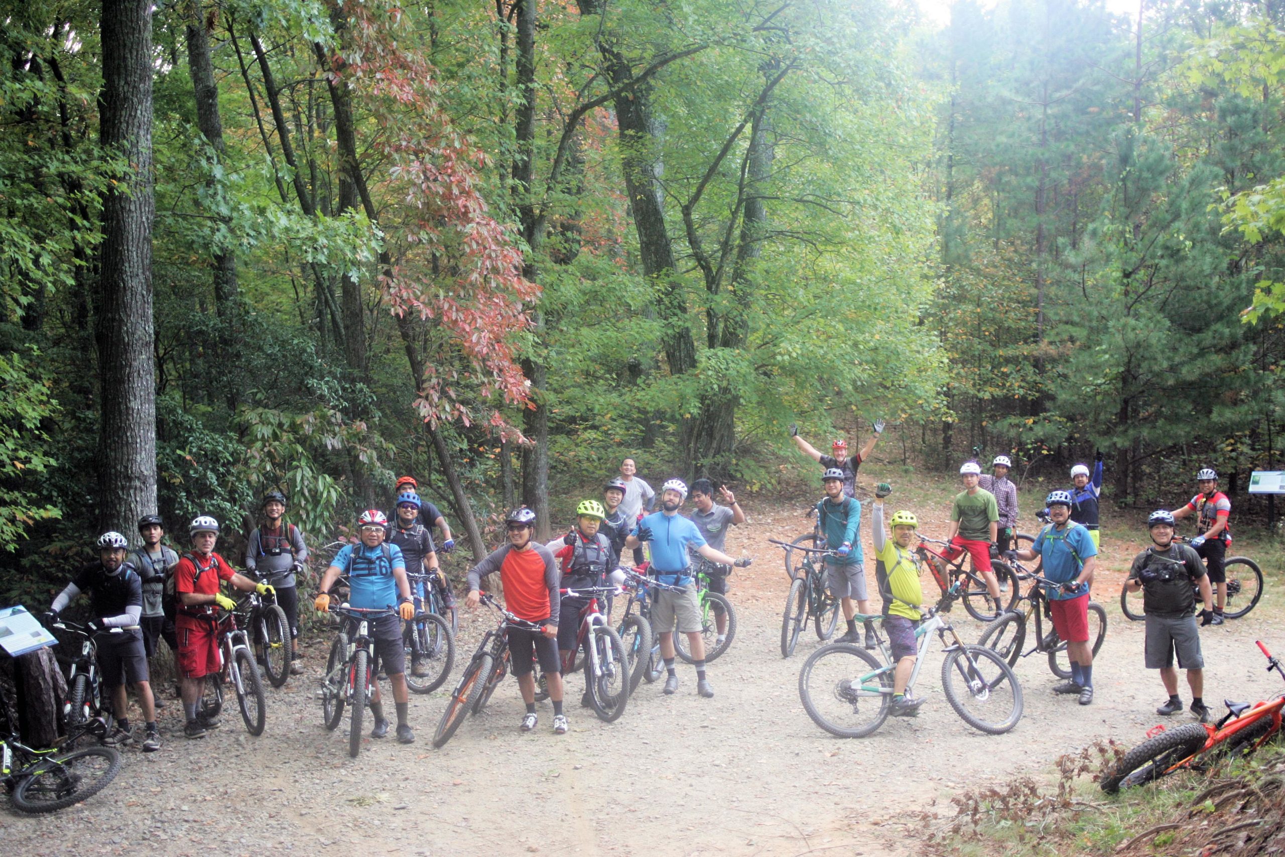 A group of mountain bikers poses for a photo on a dirt path surrounded by lush green trees. Some members are on their bikes, while others stand with their hands raised in excitement. The scene captures a sense of camaraderie and outdoor adventure, with vibrant fall foliage beginning to show through the green leaves. Bent Creek mountain bike trail.