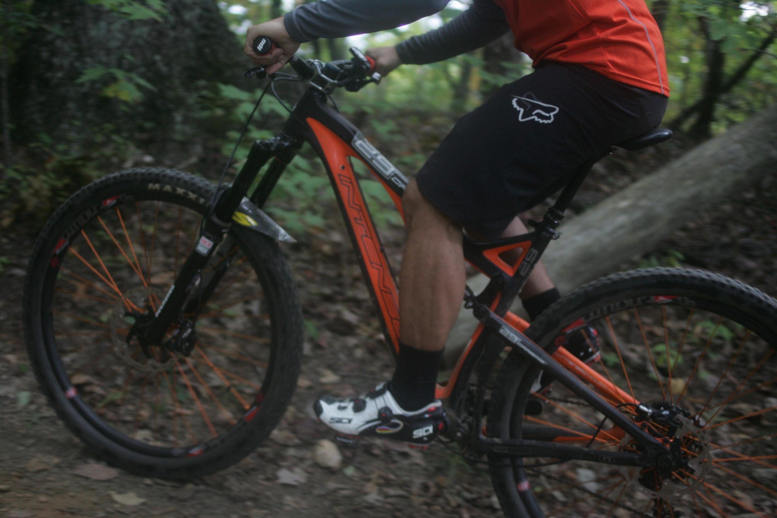 A close-up image of a cyclist riding a mountain bike on a wooded trail. The bike features a black and orange frame, and the cyclist is wearing a red shirt and black shorts. The background includes greenery and dirt, highlighting the outdoor biking environment. Bent Creek mountain bike trail.