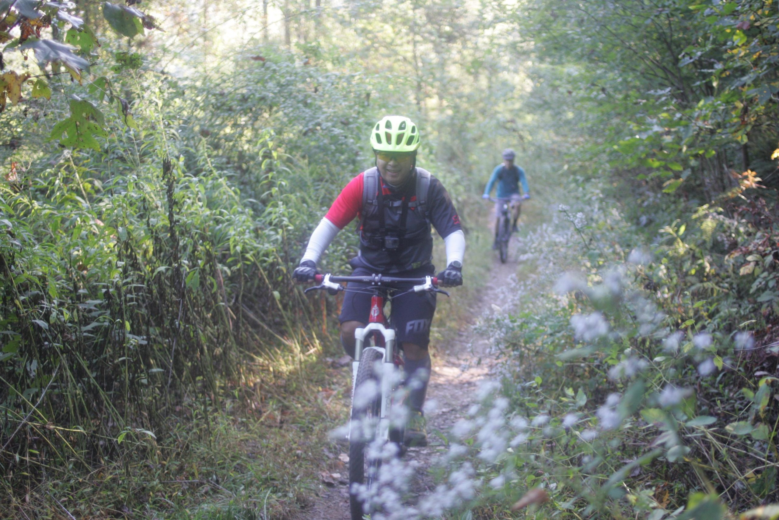 Two mountain bikers ride along a narrow trail surrounded by lush greenery. The first biker, wearing a bright yellow helmet and a red and black jersey, smiles as he pedals forward. The second biker, dressed in a teal shirt, follows behind on the same path. Sunlight filters through the trees, illuminating the scene and creating a vibrant outdoor atmosphere. Bent Creek mountain bike trail.