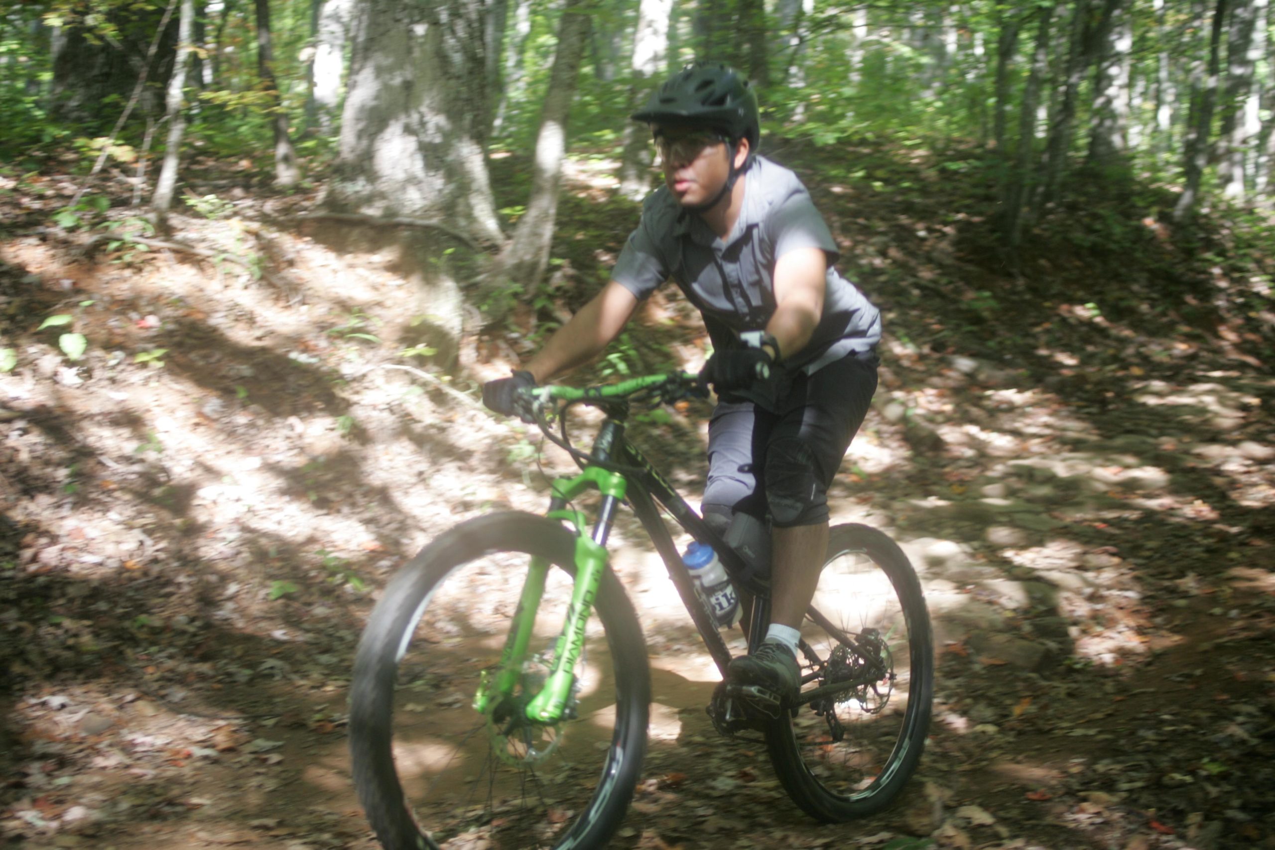 A person riding a mountain bike on a dirt trail surrounded by trees, wearing a helmet and gloves. The scene is vibrant with sunlight filtering through the foliage, and the rider appears to be navigating through a wooded area with fallen leaves on the ground. Bent Creek mountain bike trail.