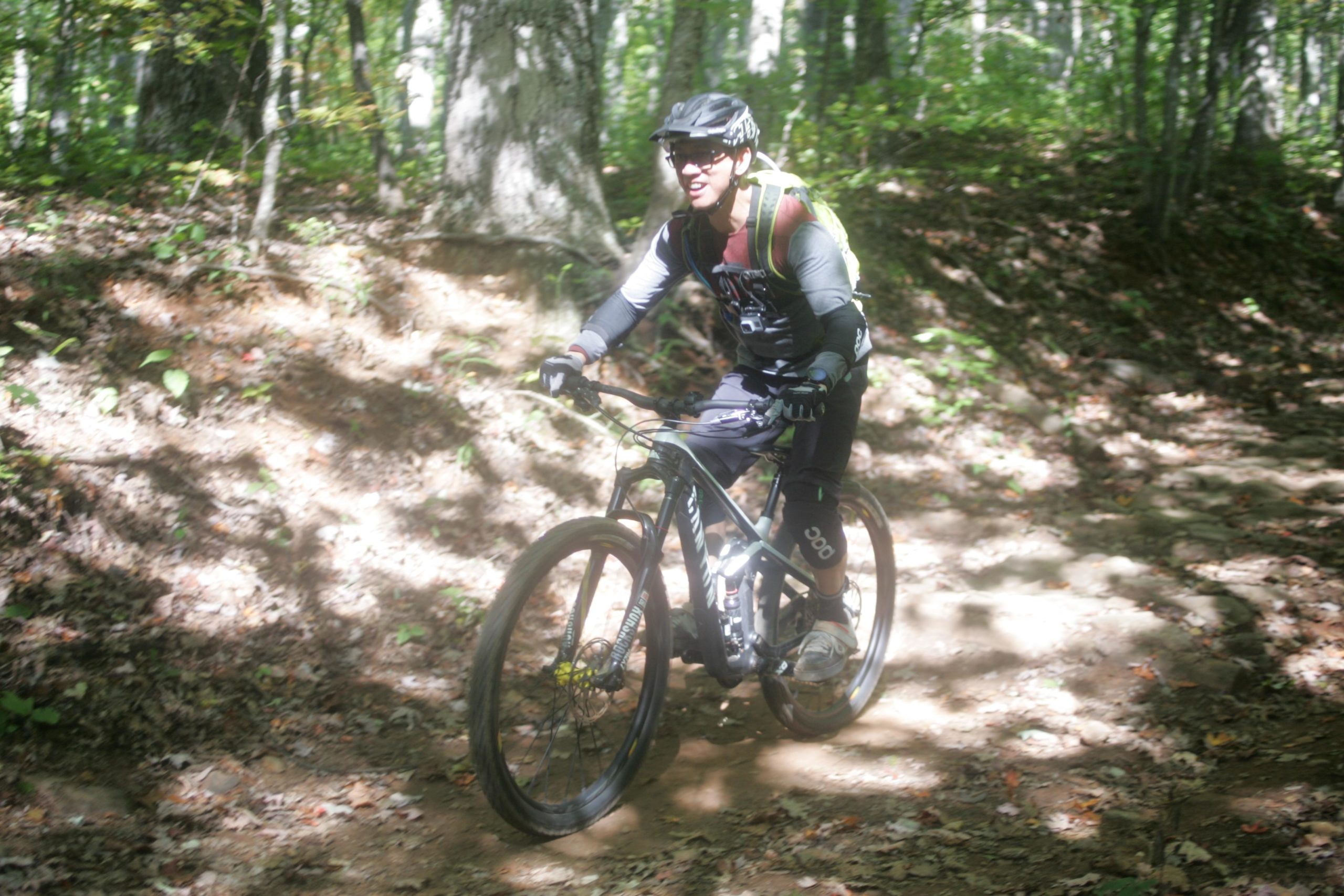 A cyclist riding a mountain bike on a dirt trail through a forest, surrounded by trees and fallen leaves. The rider is wearing a helmet, sunglasses, and cycling gear, appearing focused and enjoying the ride on a sunny day. Bent Creek mountain bike trail.