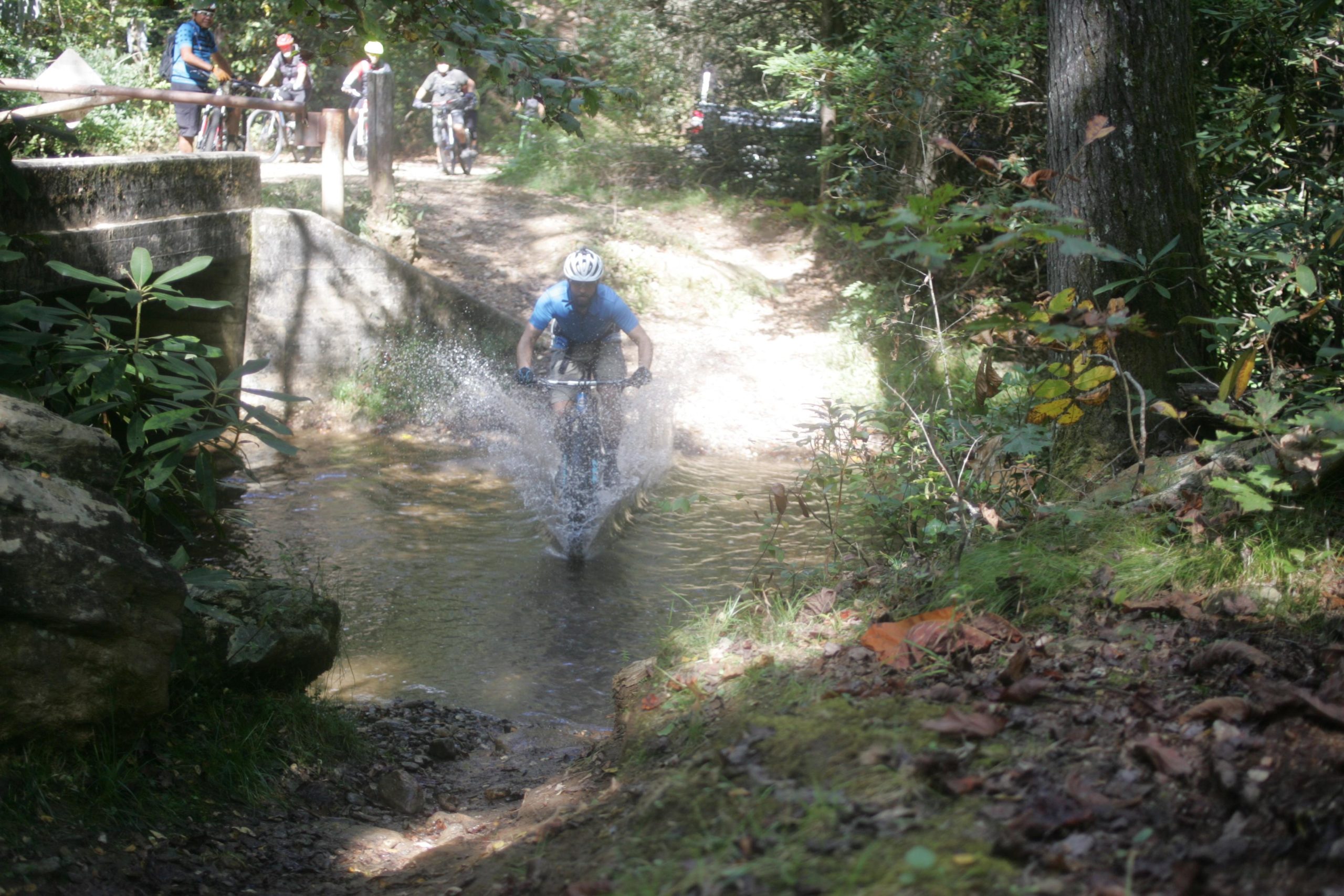 A mountain biker riding through a shallow stream, splashing water around, with a group of cyclists in the background on a forest trail surrounded by greenery. Bent Creek mountain bike trail.